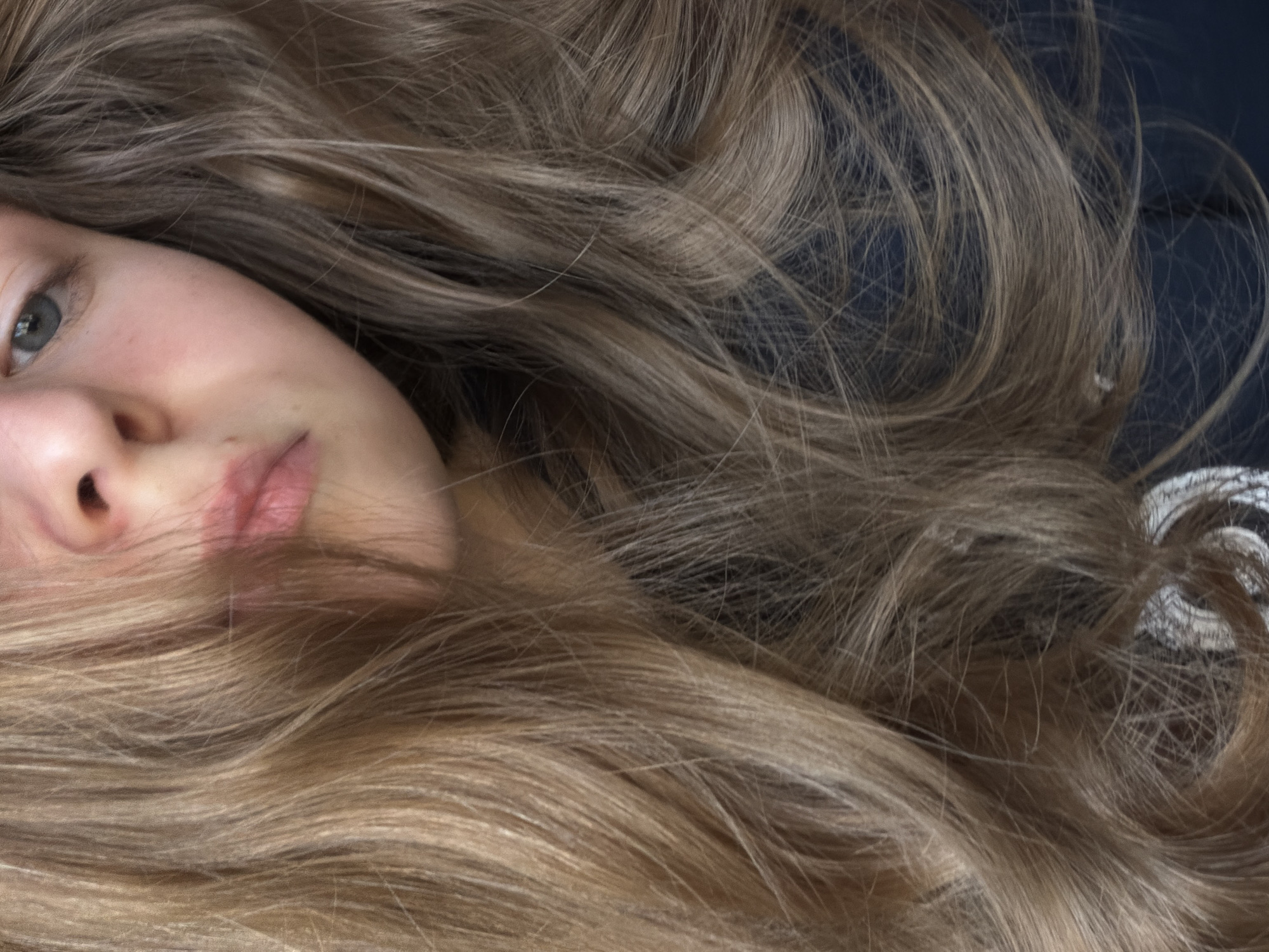 A close-up portrait of a young woman with light brown, shoulder-length hair spread around her face. Her face is turned slightly towards the left, revealing one grey eye and a small section of her nose and lips. The background is out of focus, but appears to be a dark blue or grey color. There is a small section of what looks like fabric with a swirl pattern towards the top right. The location is not discernible from the visual content.