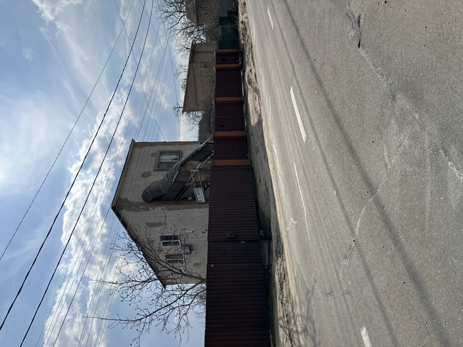 The image shows a residential street in Horenka, Ukraine (latitude: 50.525, longitude: 30.241) with a two-story house on the left side of the street. The house is damaged and appears to be in the process of being repaired, as scaffolding is visible. There is a brown fence in front of the house, and a second house is visible in the background. The street is paved with asphalt and has white markings for traffic lanes. The sky is partly cloudy with some sunlight shining through. The scene is empty of people.