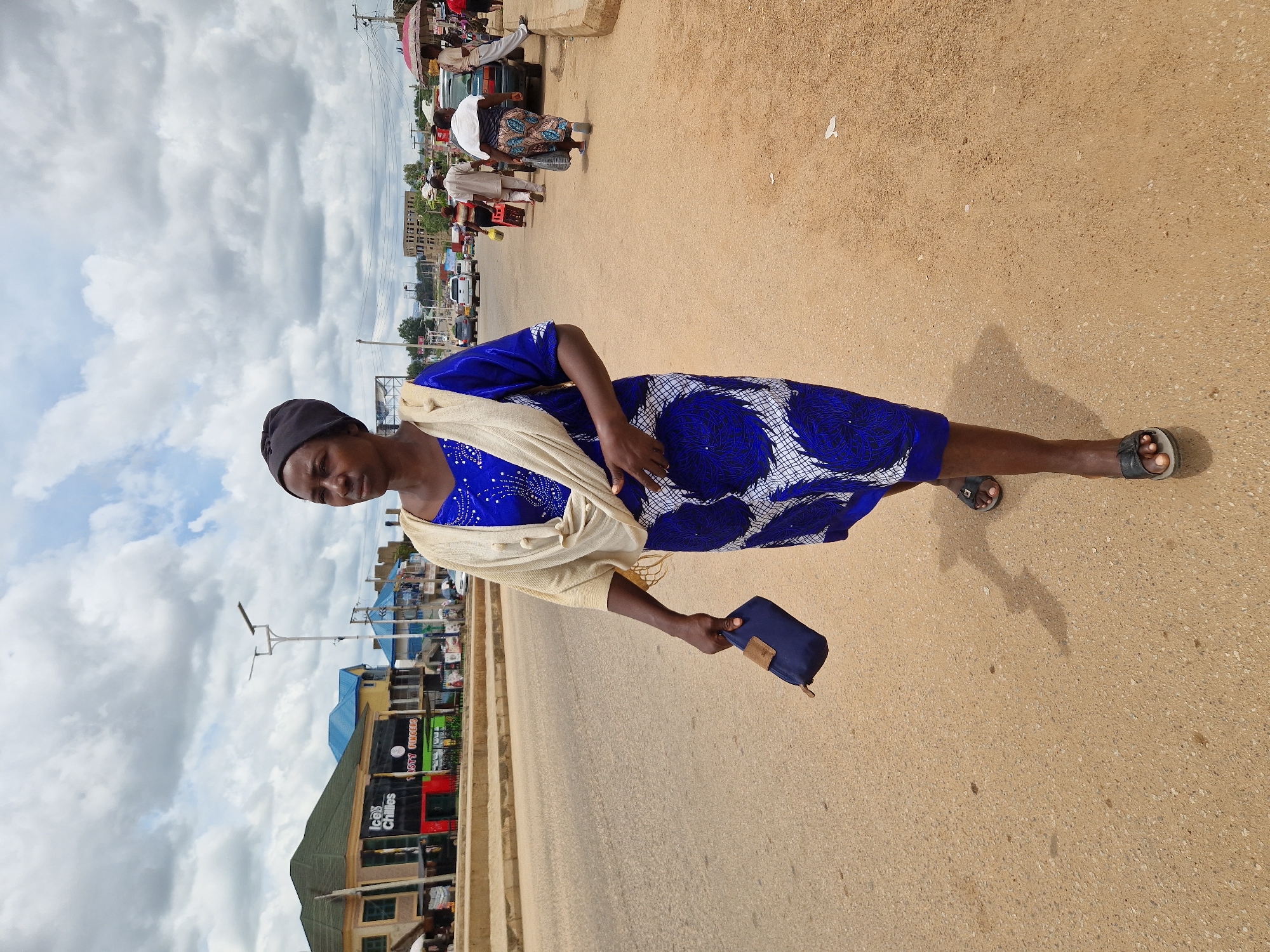 A woman walks on a dusty road in Kabong, Nigeria, with a blue and white patterned dress, a beige shawl, and a black headscarf. She is carrying a dark blue pouch in her right hand and wearing black sandals. In the background, buildings line the street, with some advertising banners. Several other people are walking and standing along the road, and vehicles are present. The sky is cloudy.