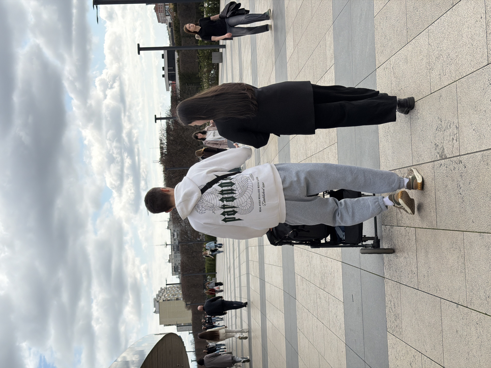 The image shows a group of people walking on a paved area in what appears to be a city park or public space. In the foreground, a person in a white hoodie and grey sweatpants is either sitting in or pushing a wheelchair. Behind them, a woman in a black coat is walking. Further back, other people are visible, along with buildings and trees. The sky is overcast with many clouds.
Based on the architectural style of the buildings and the general environment, the location is likely not in Krasnodar, Russia. The buildings are European in style.