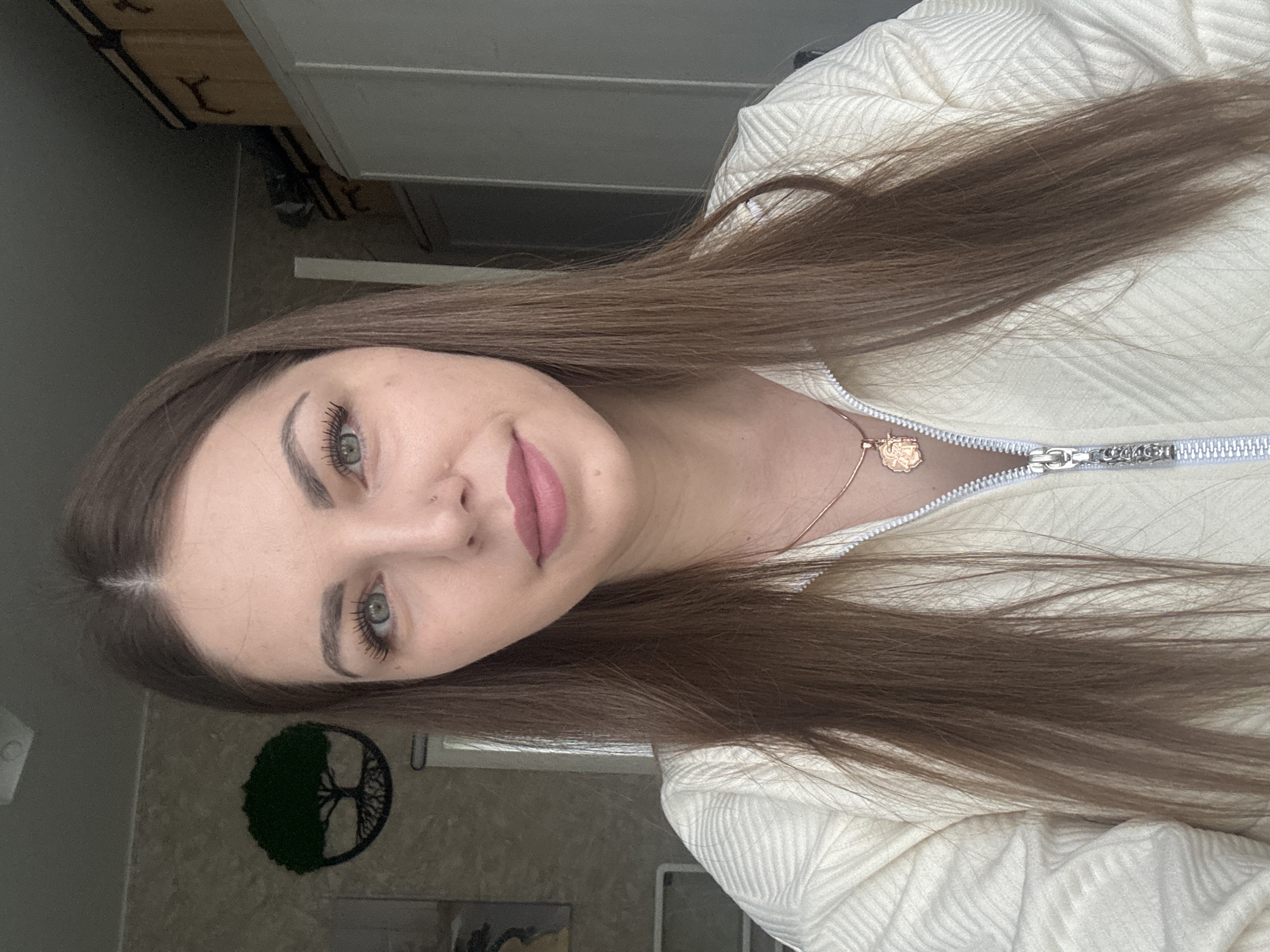 A young woman with long, light brown hair is centered in the frame, tilting her head towards the camera. She has fair skin, light green eyes, and pink lipstick. She's wearing a light-colored, textured sweater with a zipper. A gold necklace with a charm is visible on her neck. The background includes a wall with a tree-of-life decoration, a portion of a cabinet, and other indistinct items suggesting an interior space. The image does not provide evidence of the location being in Раздолье, Russia.