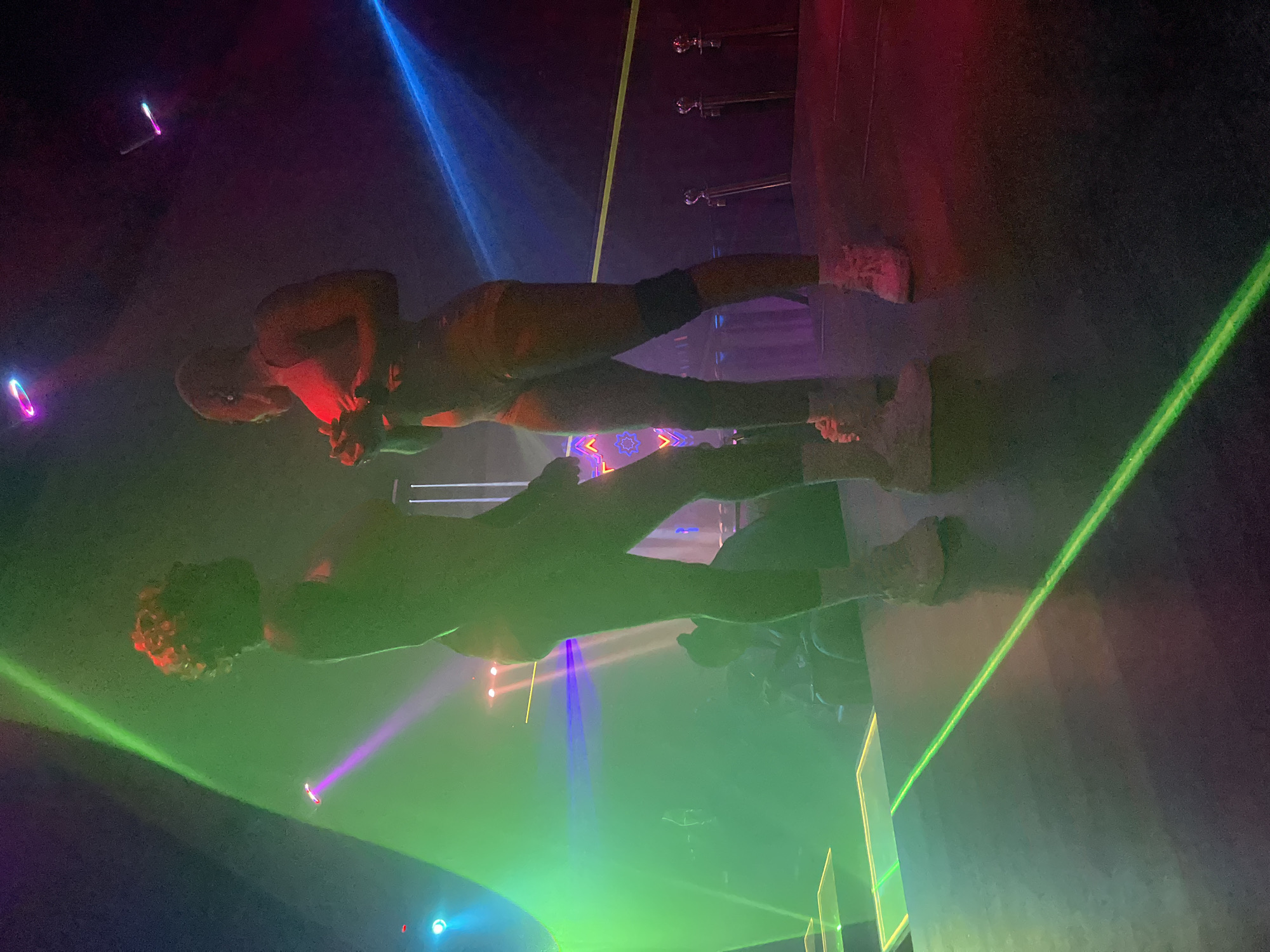 The image depicts a dimly lit indoor stage with vibrant stage lighting, including green, purple, and blue beams. Two individuals are standing on the stage, silhouetted against the lights. The person on the left has curly hair and is facing away from the camera. The person on the right is facing left, towards the other individual, and appears to be holding something in their hands. Both individuals are wearing athletic-inspired outfits and sneakers. The stage itself has reflective surfaces, and a neon green laser line is visible across the floor. In the background, there are glimpses of an audience and stage rigging. The overall atmosphere suggests a performance or event. The location is stated to be Howayi, Nigeria.
