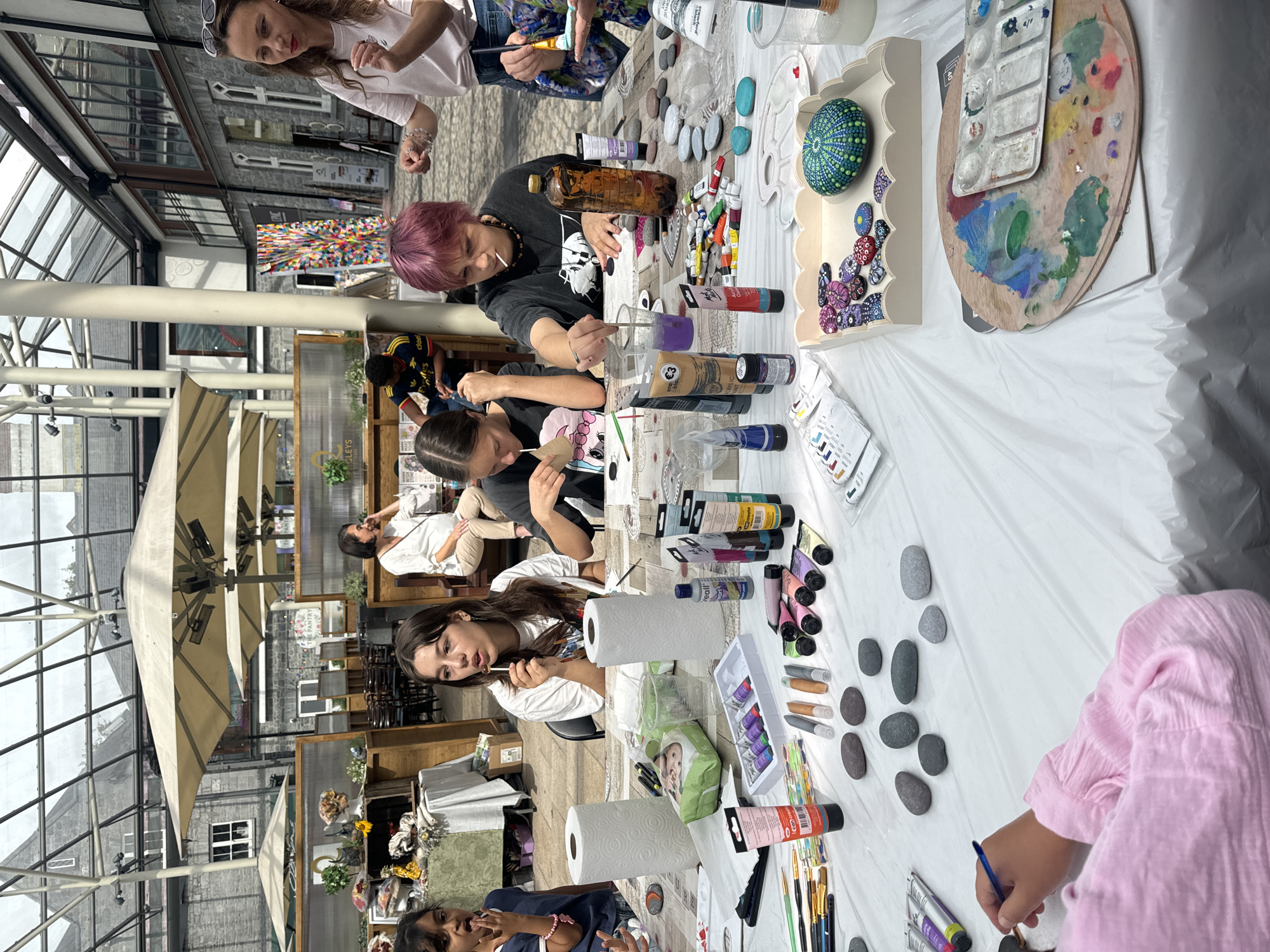 This image shows an art workshop in Kilkenny, Ireland, likely at the Kilkenny Design Centre. A group of people, primarily young women and children, are seated at a table covered with a white plastic sheet, engaged in painting pebbles. Various art supplies are spread across the table, including tubes of paint, brushes, water cups, rolls of paper towels, and palettes. In the foreground, a young person in a pink top is holding a paintbrush, actively painting a pebble. Other individuals are seen with paintbrushes near their faces, indicating they are also painting. In the background, there are other tables with displays and people in the background, suggesting a public event or market setting. The environment appears to be an indoor area with a glass roof, allowing natural light to illuminate the scene, with architectural elements characteristic of the Kilkenny Design Centre visible.