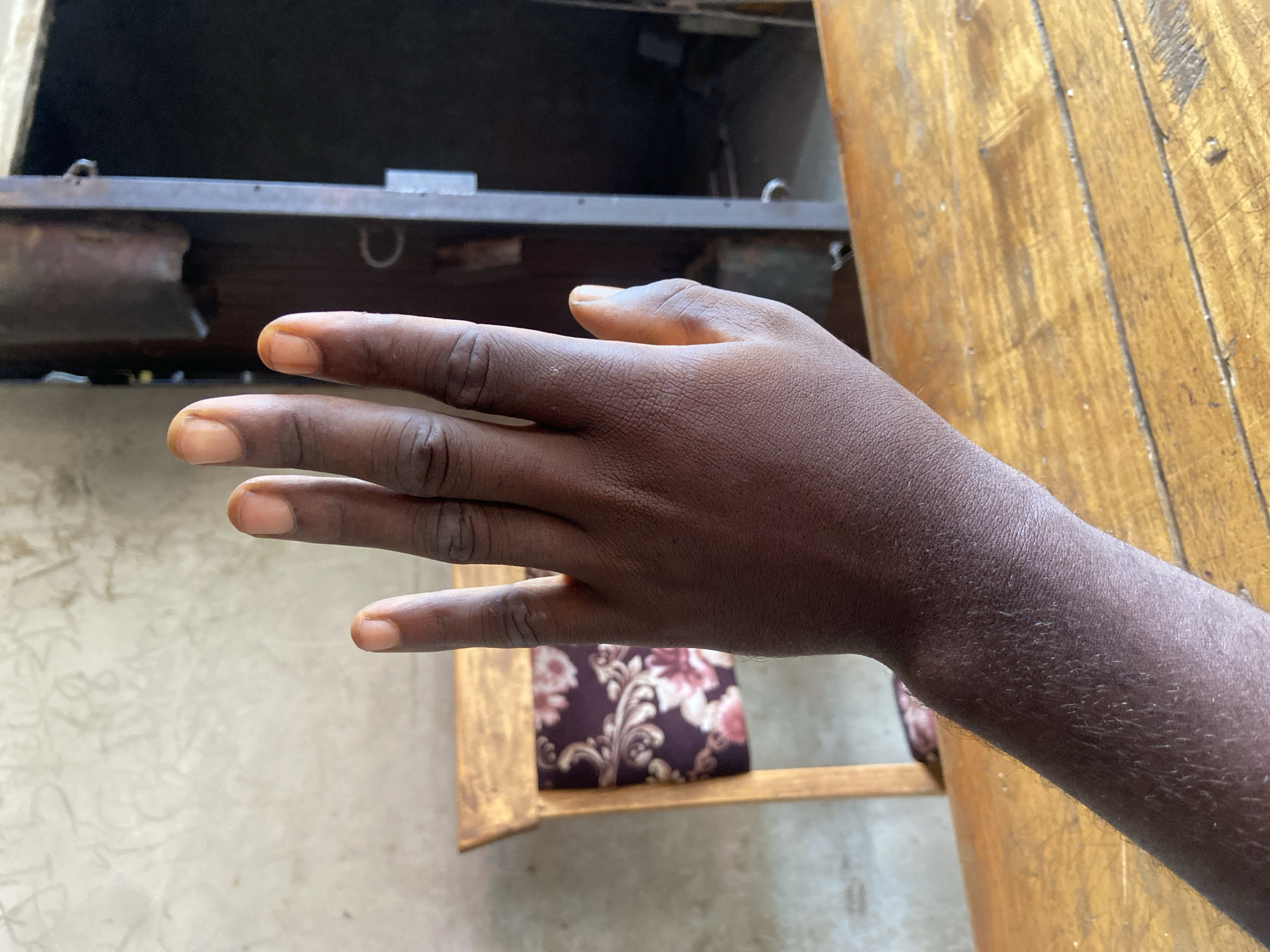 A close-up photograph shows a dark-skinned human hand and arm extending from the lower right towards the left. The fingers are outstretched. The setting is indoors. The background includes a light-colored wall or surface, a wooden table to the right, and what appears to be a metal structure. Below the hand, a chair with a floral-patterned cushion is visible. The location is Mararaba, Nigeria.
