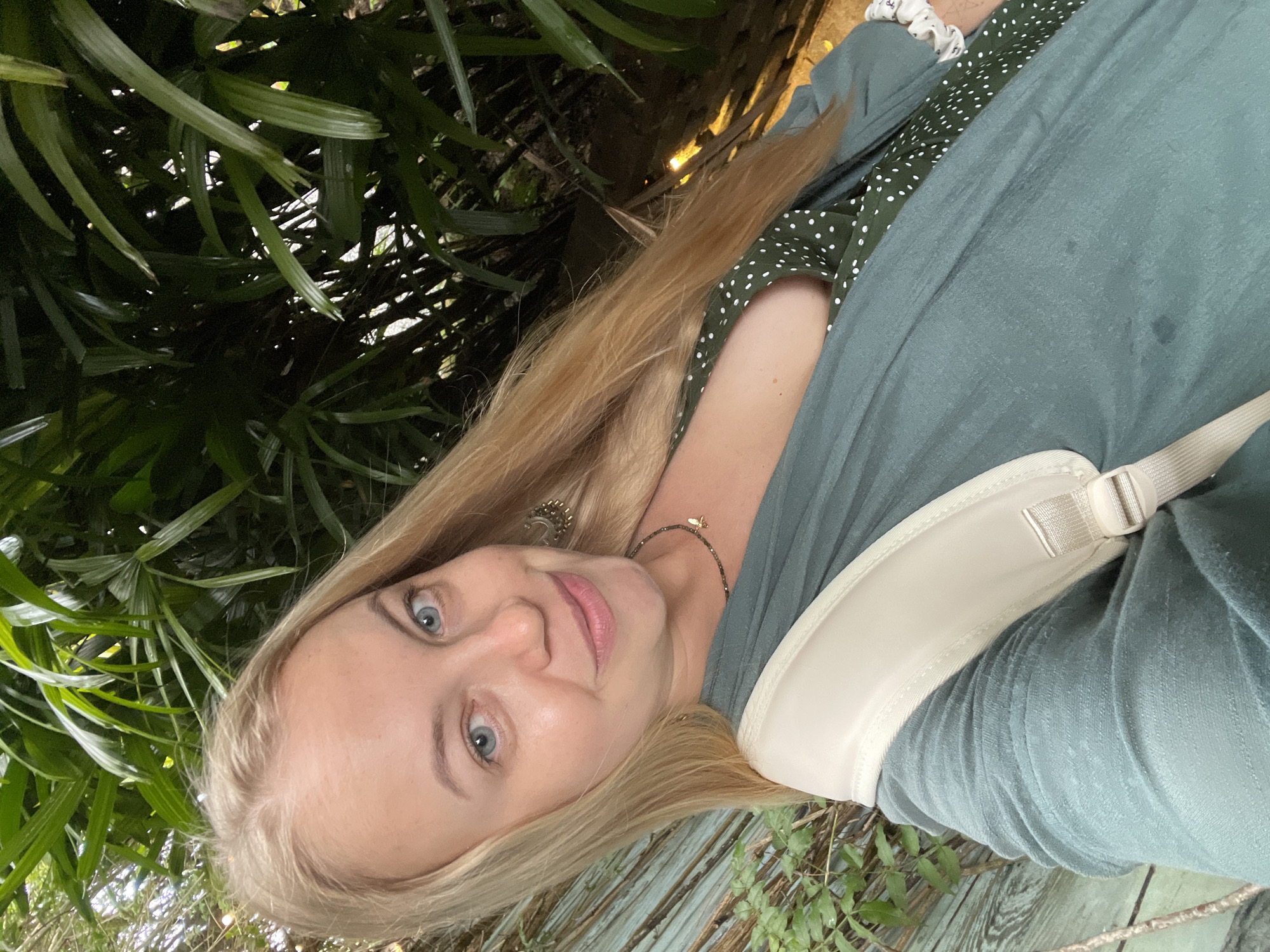 A blonde woman smiles at the camera, taking a selfie. She is outdoors, with lush green foliage on the right side of the frame and a wooden fence in the background on the left. She wears a light green open-front cardigan over a dark green polka-dot dress. A cream-colored bag strap is visible over her shoulder. Her hair is long and blonde. She is wearing a necklace with a small gold charm. The environment suggests a tropical or subtropical setting. The location is identified as Da Lat, Vietnam, a city known for its cooler climate and lush vegetation.