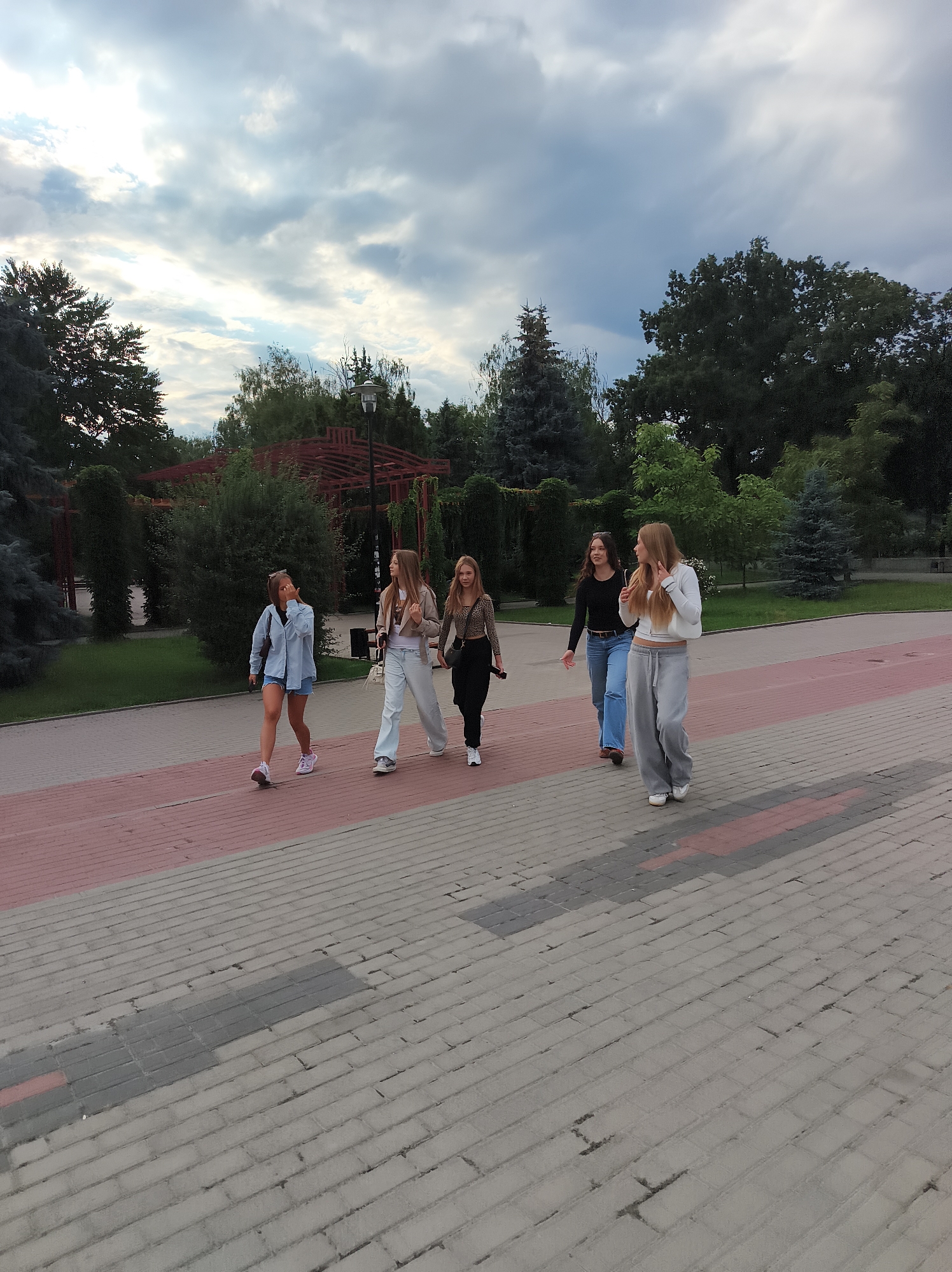 In Ternopil, Ukraine, five young women walk on a paved pathway in a park. The pathway is made of light grey and dark grey bricks, with a red brick border. The women are dressed in casual, modern clothing. The woman on the far left wears a light blue denim jacket and shorts, with white sneakers. Next to her, a woman in a white shirt and white pants walks with her hand near her face. The third woman from the left has on a black top and blue jeans. The fourth woman wears a white hoodie and grey sweatpants. The woman on the far right is dressed in a black and leopard-print top with black pants and white sneakers. The park features lush green trees and bushes, with a wooden structure visible in the background, possibly a gazebo or pavilion. The sky is overcast with dramatic clouds.