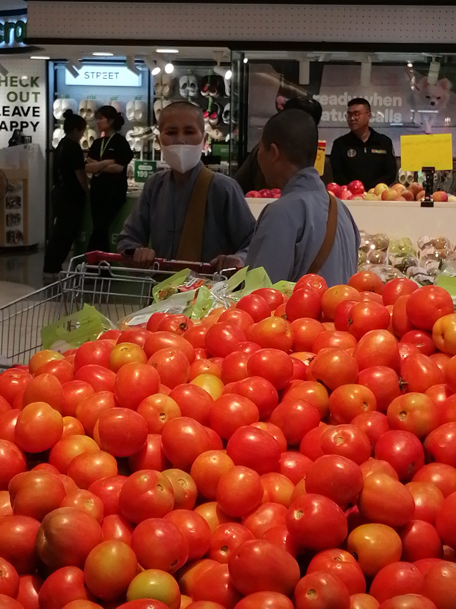 The image appears to be taken inside a supermarket or a large retail store. The foreground is dominated by a large pile of red tomatoes. Behind the tomatoes, a shopping cart is partially visible. In the middle ground, two individuals wearing monastic-like robes are standing, one of whom is wearing a white face mask. Behind them, there are shelves stocked with various items, including footwear, and a sign that says "STREET". Further in the background, a man in a dark uniform, possibly a security guard, is visible. The overall lighting suggests an indoor, well-lit environment. There is no visual information within the image to suggest the location is specifically Da Lat, Vietnam.