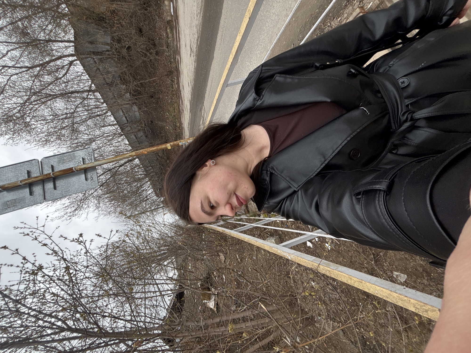 This outdoor portrait features a young woman in what appears to be an urban or semi-urban setting in Perm, Russia, during late winter or early spring. The image is composed at a Dutch angle, tilting the horizon.
The main subject is a woman with dark hair, visible from her mid-torso upwards. She is dressed in a black, possibly faux leather, long-sleeved coat that is tied at the waist, worn over a dark brown, scoop-neck top. A small, circular, silver-colored stud earring is visible in her left ear. She looks slightly to her left with a gentle, pleasant smile.
The background indicates an overcast day, consistent with the bare branches of trees that show nascent buds, suggesting the very beginning of spring. Behind the woman, a light-colored road or path with a prominent yellow dividing line is visible, bordered by a metal barrier or railing. On a rusty metal bar, faint Cyrillic letters that resemble "ПО" (PO) can be discerned. Further into the background, a silver construction ladder lies on the ground amidst dry, sparse vegetation and dirt. The overall scene conveys a cool, transitional seasonal atmosphere in a casual outdoor environment.