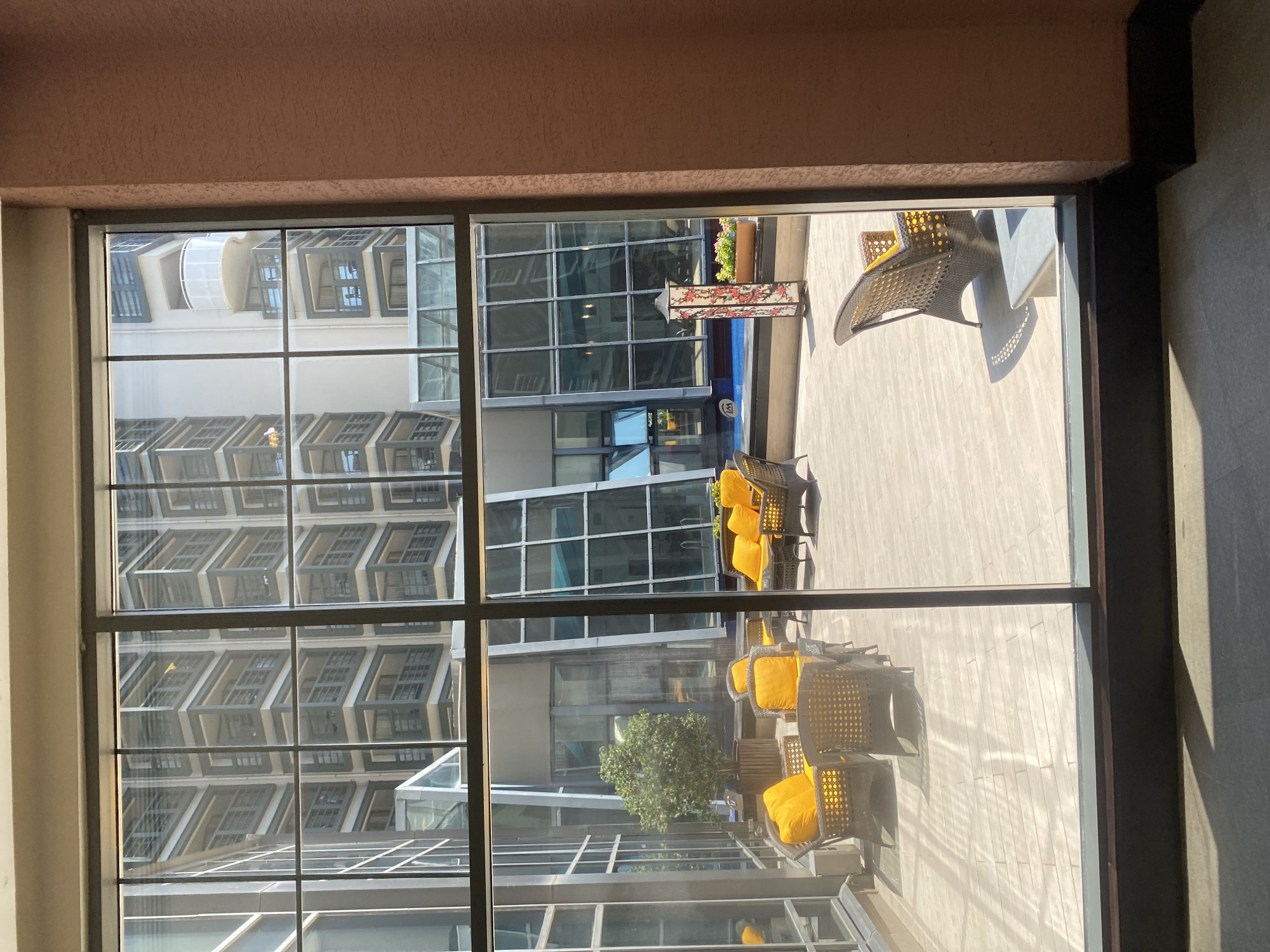 This is an exterior shot of a courtyard taken from inside a building through a large window. The courtyard is paved with light-colored tiles and features several pieces of outdoor furniture, including wicker chairs with yellow cushions and a wicker sofa. A small tree in a planter is also present. In the background, a swimming pool with blue water is visible, along with a decorative lamp and a planter box with red flowers. Beyond the courtyard, modern buildings with geometric designs and balconies are reflected in the glass walls. The overall impression is of an urban, resort-like setting. The image does not provide enough visual information to definitively identify the location as Aksu, Turkey.