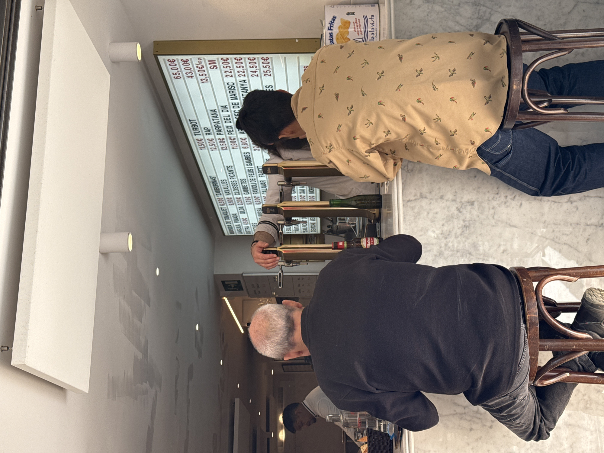 The image shows the interior of a bar or restaurant, possibly in Barcelona, Spain, based on the Spanish menu board in the background. Two men are seated at the bar, one wearing a black sweater and grey jeans, the other wearing a light tan shirt with a bird pattern and dark blue jeans. The man in the black sweater is seated to the left and is partly bald with grey hair, while the other man has dark hair. A bartender or server is seen reaching across the bar to serve a drink. The menu board shows prices in euros for various seafood dishes like turbot, rap, parpatana, and gambetes. Brands of beer, likely Spanish, are displayed, including Mahou. A small box of "Latas Fritas" (fried canned goods) is visible. The general atmosphere appears casual and social, capturing a typical bar scene.