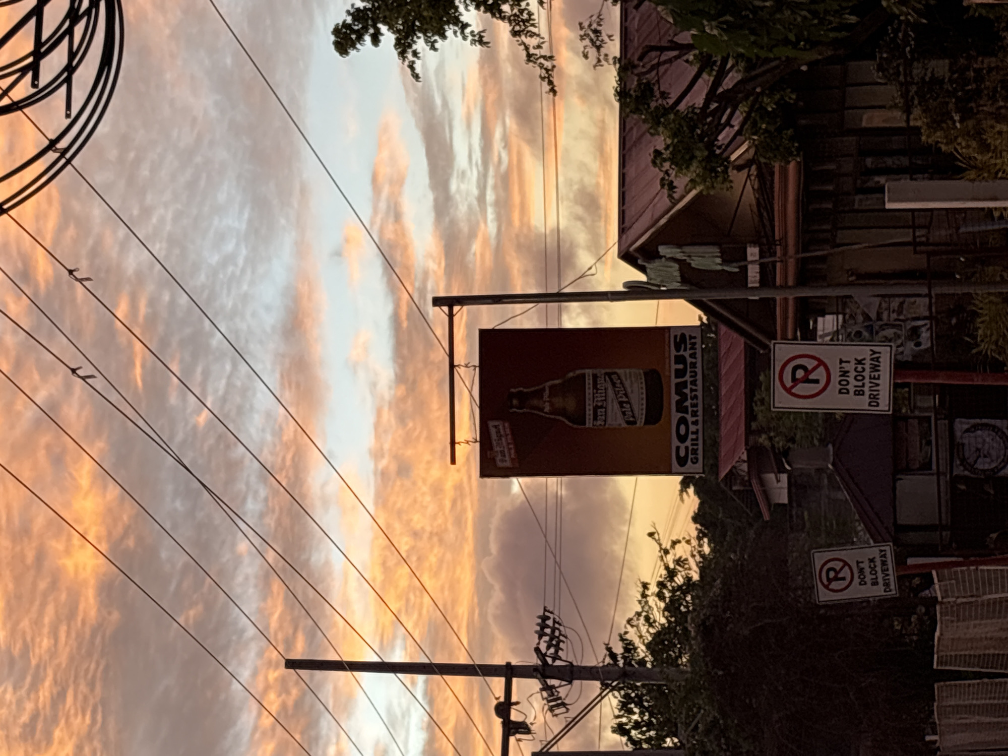 The image captures a sunset scene in Davao City, Philippines, with the sky filled with hues of orange and pink clouds. In the foreground, a sign for "COMUS GRILL & RESTAURANT" is prominently displayed, featuring an advertisement for San Miguel Pale Pilsen. Two "DON'T BLOCK DRIVEWAY" signs are visible on the left and right sides of the frame. Utility poles and electrical wires crisscross the sky, indicating an urban or suburban setting. The background reveals a glimpse of a building with a reddish roof and lush green trees, typical of the tropical environment of Davao City. The overall ambiance is serene, with the natural beauty of the sunset contrasting with the man-made structures.