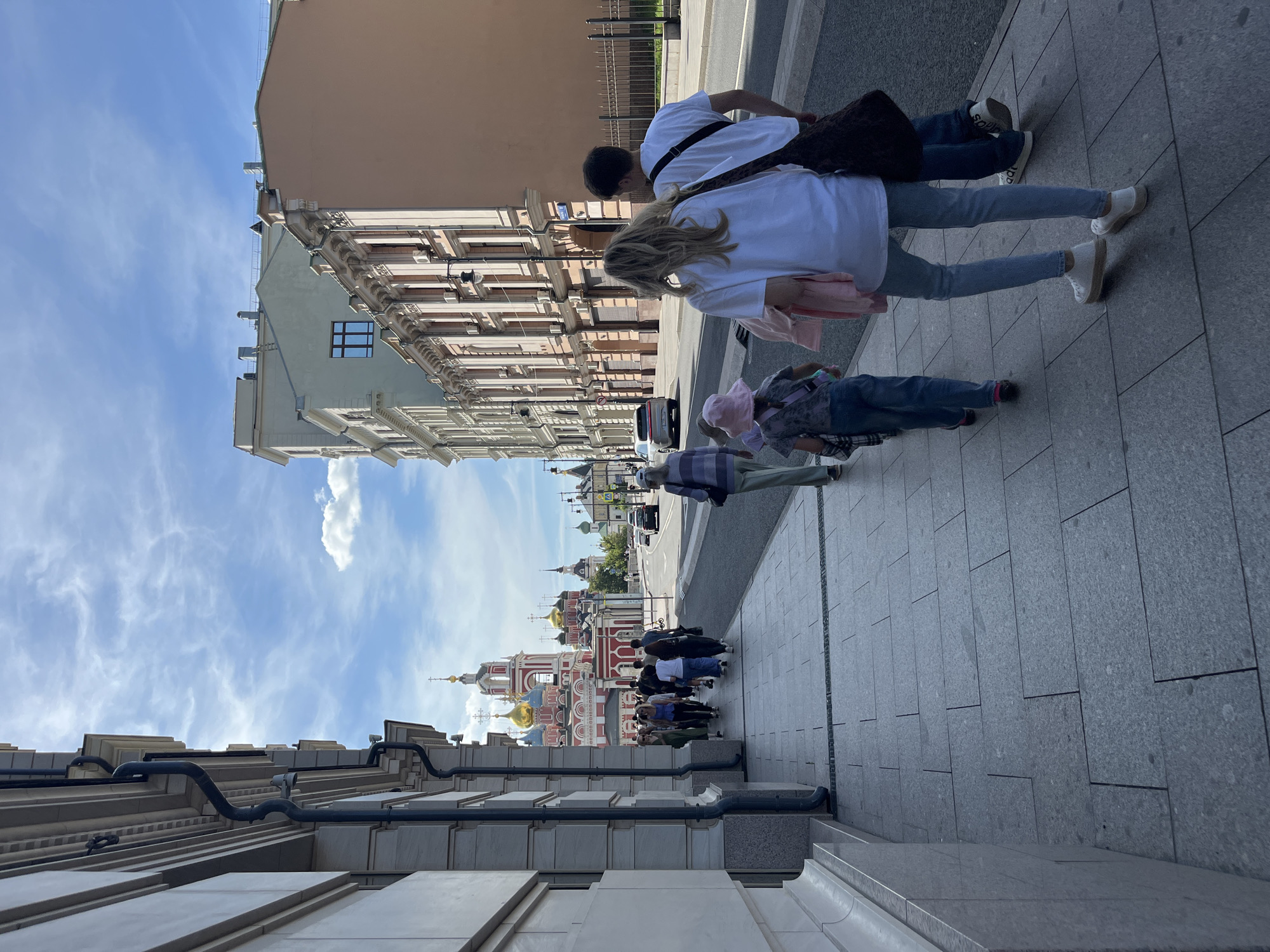 A group of people walk along a sidewalk on a sunny day in Moscow, Russia. The sidewalk is paved with gray square tiles. On the left side of the image is a light-colored building with ornate architectural details and dark downspouts. On the right side of the image, there are several buildings with light brown and cream facades, featuring multiple windows and air conditioning units. In the distance, a red and white church with golden domes is visible. The street is paved with asphalt and has cars driving on it. The sky is blue with scattered white clouds. Several individuals are walking in the same direction on the sidewalk, including a man and a woman in the foreground, a child wearing a pink hat, and another child in the middle ground. A few other people are walking ahead of them, further down the sidewalk.