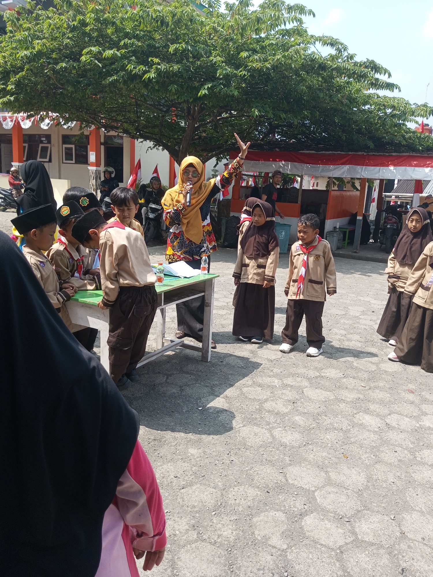 The image depicts an outdoor gathering in the city of Kalibeber, Indonesia, likely a school event given the uniforms worn by the children. In the center, a woman in a yellow hijab and a vibrantly patterned top stands, speaking into a microphone and gesturing with her left hand. She appears to be addressing a group of children, some of whom are wearing brown scout uniforms with red and white neckerchiefs, and others in black caps.
In the foreground and mid-ground, several children are visible. Some are looking towards the speaker, while others are engaged with a green table where books and bottles of water are placed. The ground is paved with hexagonal concrete blocks. Behind the gathering, there is a building with orange and white trim, adorned with red and white flags, suggesting a festive occasion or national celebration. A large tree with lush green leaves provides shade over the scene. Several motorcycles are parked in the background, indicating the mode of transportation used by some attendees. The overall atmosphere appears to be a daytime educational or community event.