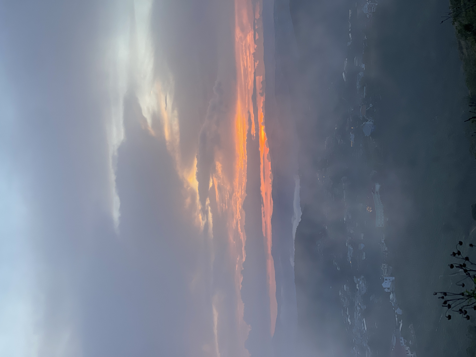 The image depicts a sunset view over a misty landscape. The sky transitions from blue to orange with heavy cloud cover. Below the horizon line, houses are clustered in a valley or on a hillside, partially obscured by mist. Vegetation is visible on the right. No people are visible in the image. While I cannot ascertain the location in Trường Thọ, Vietnam, the photo appears to be taken from a high vantage point.
