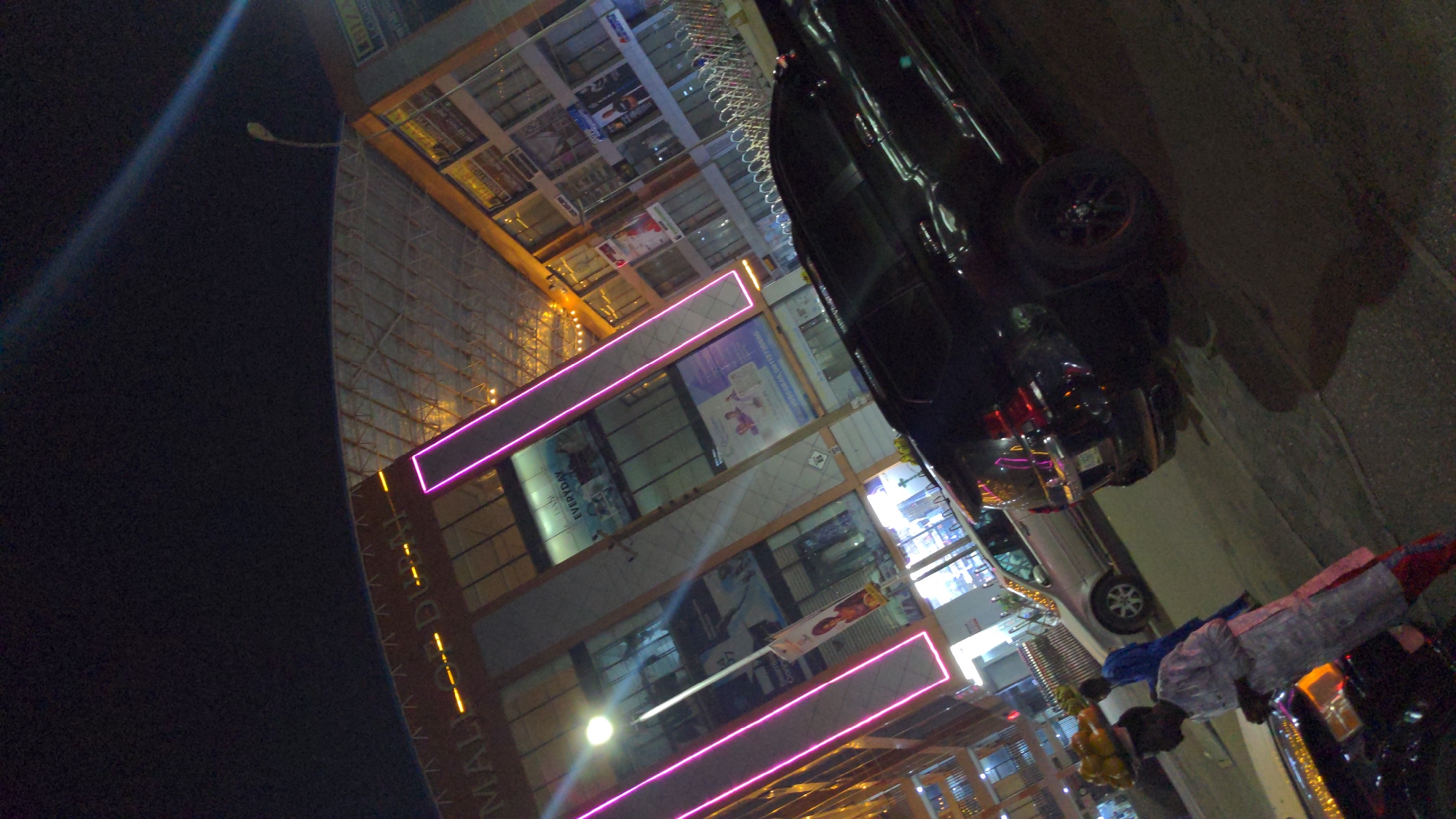 This is a nighttime photograph depicting a busy street scene in what appears to be a commercial area. The image is taken from a slightly upward angle, showing a multi-story building on the left and a road with parked cars and people on the right.
The most prominent subject on the left is a modern building, likely a mall or commercial complex, illuminated by artificial light. Its facade features large glass windows, reflecting ambient light. Striking pink neon light strips run horizontally across different levels of the building, creating a vibrant glow. Partially visible yellow vertical lights on the building spell out "MALL". Inside the building windows, various advertisements and signs are visible, with some text vaguely decipherable as "EVERYDAY". Scaffolding is present on the upper left section of the building, suggesting construction or maintenance. A streetlight extends from the dark sky in the upper left corner.
On the right side of the image, a dark asphalt road is visible, with several vehicles. A large, dark-colored SUV or truck is parked prominently in the foreground, showing reflections of the neon lights. Further back, a silver car is also parked. Two individuals are standing on the right side of the road, dressed in what appears to be traditional attire; one is in blue and the other in white or light grey. They are positioned near a makeshift display of fruits, likely a street vendor's stall, with yellow and orange fruits (possibly oranges or mandarins) piled in a container.
The scene is clearly at night, indicated by the dark sky and the reliance on artificial lighting from the building, neon signs, streetlights, and vehicle lights. The overall atmosphere suggests a lively urban or commercial environment after dark.