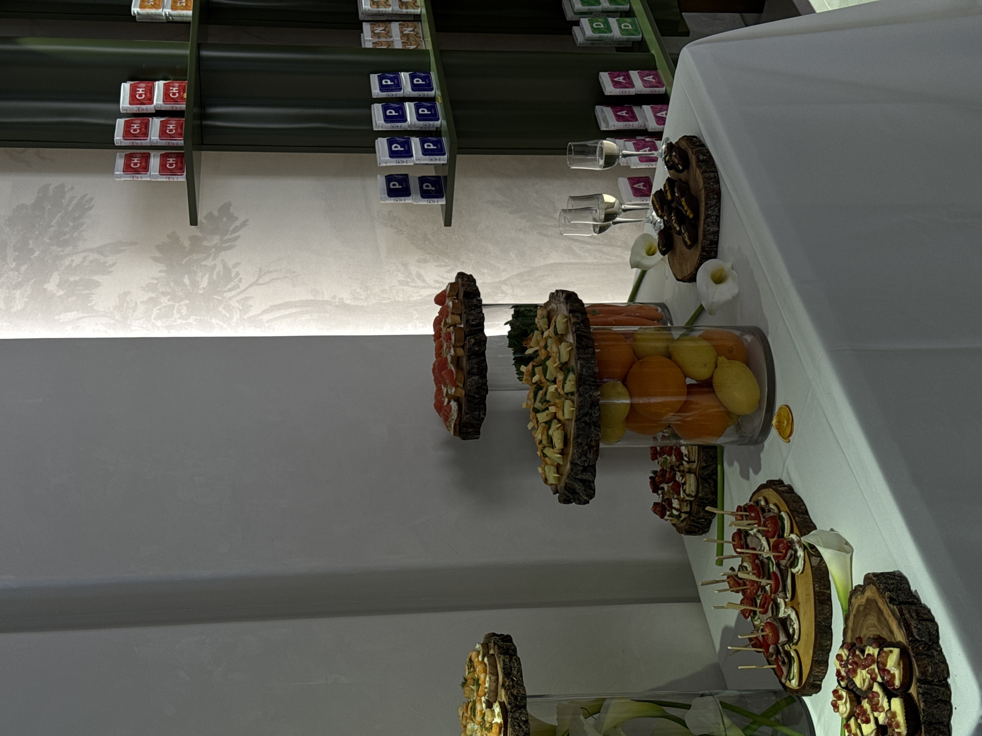 Here is a forensic description of the image:
**Scene:** The image depicts a buffet table arrangement, likely set up for an event, against a backdrop featuring shelves stocked with various packaged goods and a decorative wallpaper.
**Objects:** The table is covered with a white tablecloth. It features several serving platters arranged decoratively. These platters appear to be made of wood and display an assortment of food items, including what looks like appetizers or hors d'oeuvres arranged on skewers, slices of fruit, and pastries. A glass container holds a mix of oranges and lemons. There are also glasses, presumably for beverages, present. The shelves in the background display items labeled "CH," "P," "D," and "A" in stacks.
**Setting:** The location is not definitively identifiable as Yerevan, Armenia, from the visual information provided. However, the scene suggests a modern or upscale interior setting, potentially a hotel or banquet hall.