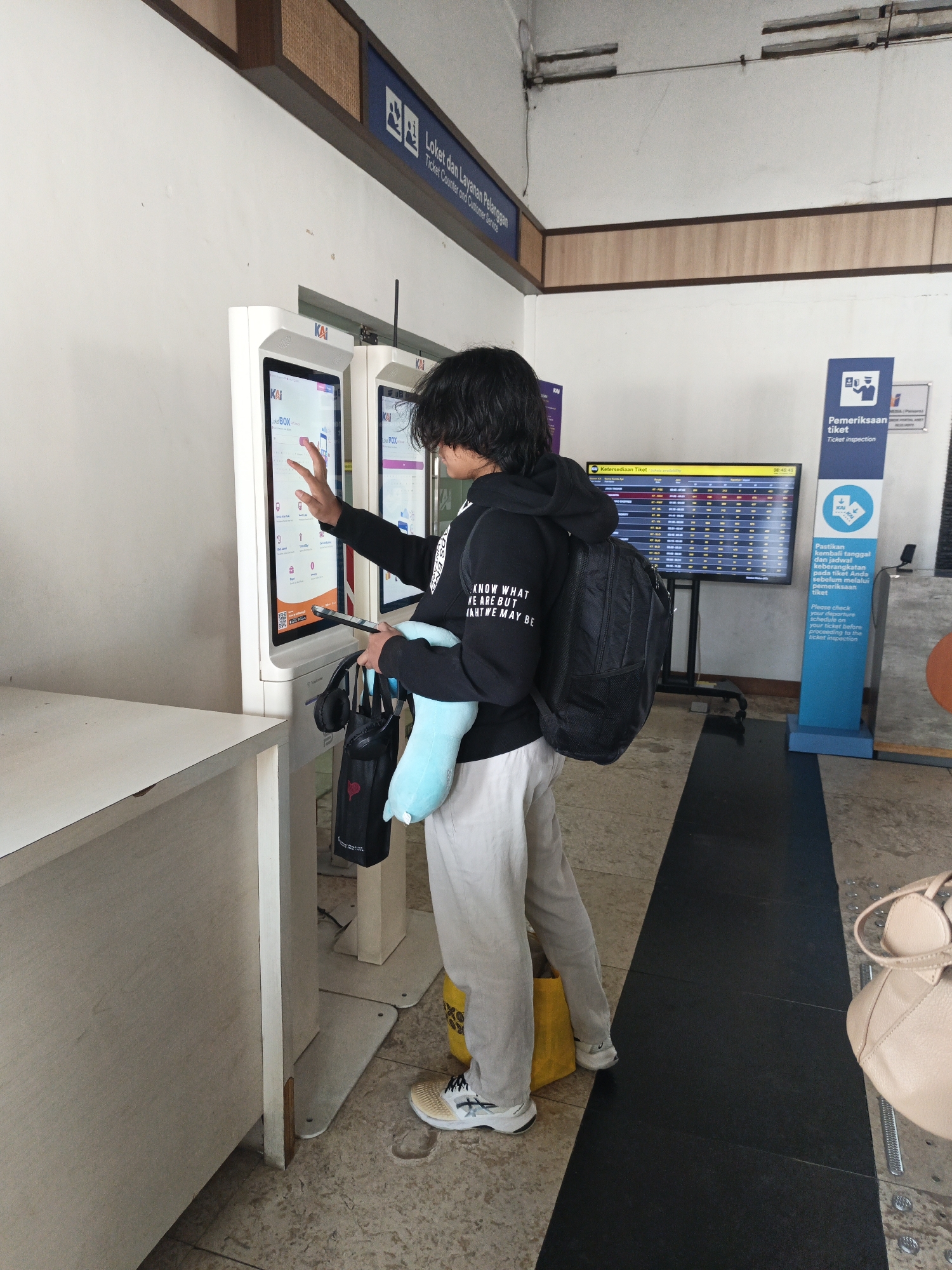 A person with long dark hair, wearing a black hoodie and light-colored pants, is standing in front of a self-service ticket kiosk at a train station in Klaten, Indonesia. The person is holding a tablet and touching the screen of the kiosk with their right hand, appearing to be interacting with it. They are also carrying a black backpack on their right shoulder and have a blue plush toy and two black bags hanging from their left arm. A large digital display screen showing train schedules is visible in the background. The environment appears to be an indoor waiting area with tiled flooring and a white wall.