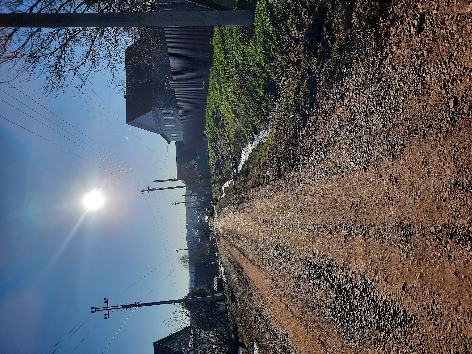 The image depicts a dirt road in Krasnogorskoye, Russia, on a sunny day. The road, composed of soil and gravel, is marked with vehicle tracks and appears somewhat muddy in places. Patches of green grass and vegetation line the side of the road, alongside houses and fences. Power lines and utility poles stretch along the road, converging in the distance. The bright sun is visible in the sky.