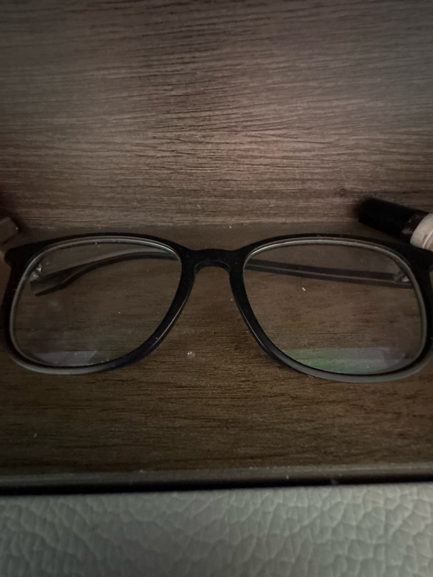 A pair of black-rimmed glasses is resting on a dark wood surface. To the right of the glasses, a black cylindrical object with a white band is visible. The background consists of the same dark wood texture. In the foreground, a textured gray surface is visible. The image is taken from a slightly elevated angle, looking down at the glasses.