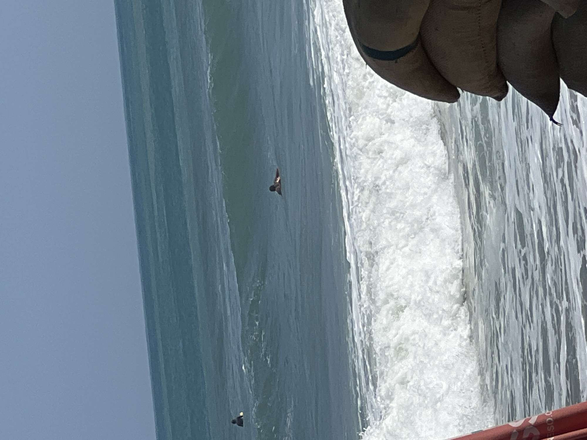 The image depicts a view of the ocean in Kuta, Indonesia, with several surfers in the water. In the foreground, a stack of sandbags is visible on the right side of the frame, partially obscuring the lower portion of the ocean. To the left, a red object, possibly a beach umbrella or surfboard, is also partially visible. The ocean itself shows moderate waves with white foam crests. Two individuals are in the water, both appearing to be surfers. One is further out in the ocean and is a small, dark figure. Closer to the foreground, another surfer is visible lying on a board, with a yellow object near them. The sky above is a clear, pale blue. The overall scene captures a typical day at the beach with recreational activities.