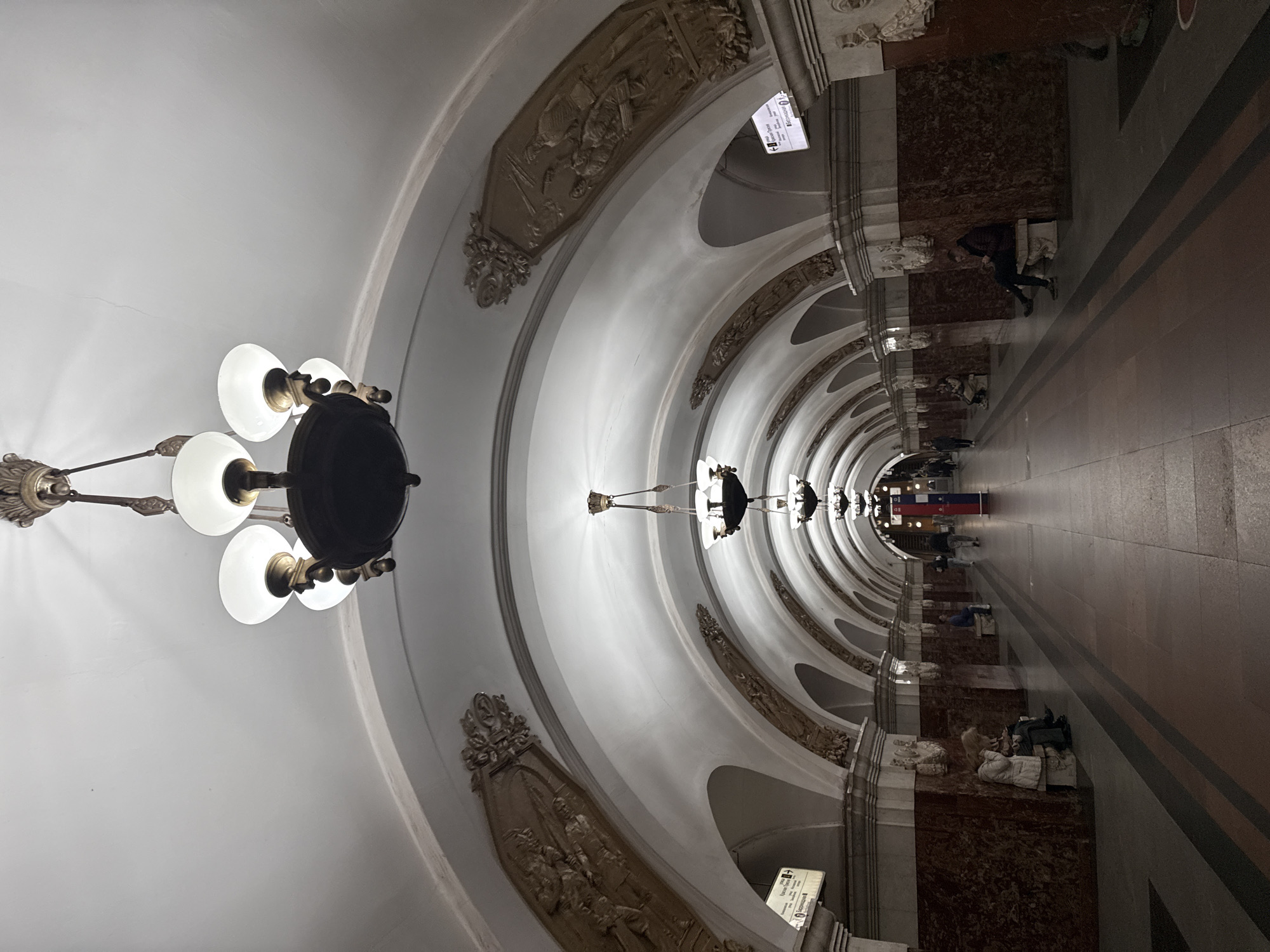 The image shows the Komsomolskaya-Koltsevaya station in Moscow, Russia. The station features a high vaulted ceiling adorned with ornate decorations and sculptures. A large chandelier hangs from the ceiling on the left side. A long hallway extends into the distance, lined with marble walls and benches. Several people are seen walking or sitting along the hallway. The overall perspective is slightly distorted, emphasizing the station's architectural grandeur.