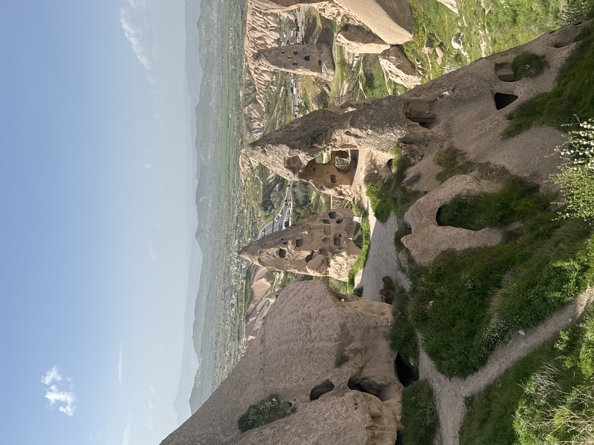 This is an outdoor shot taken in Uçhisar Beldesi, Turkey. The foreground showcases natural rock formations, some with carved-out holes resembling dwellings. Patches of green vegetation and dirt pathways are interspersed among the rocks. A road with parked cars is visible in the middle ground, nestled among the rock structures and vegetation. The background reveals a distant cityscape extending towards a mountain range, all beneath a clear, blue sky with scattered light clouds. The orientation of the image is vertical.