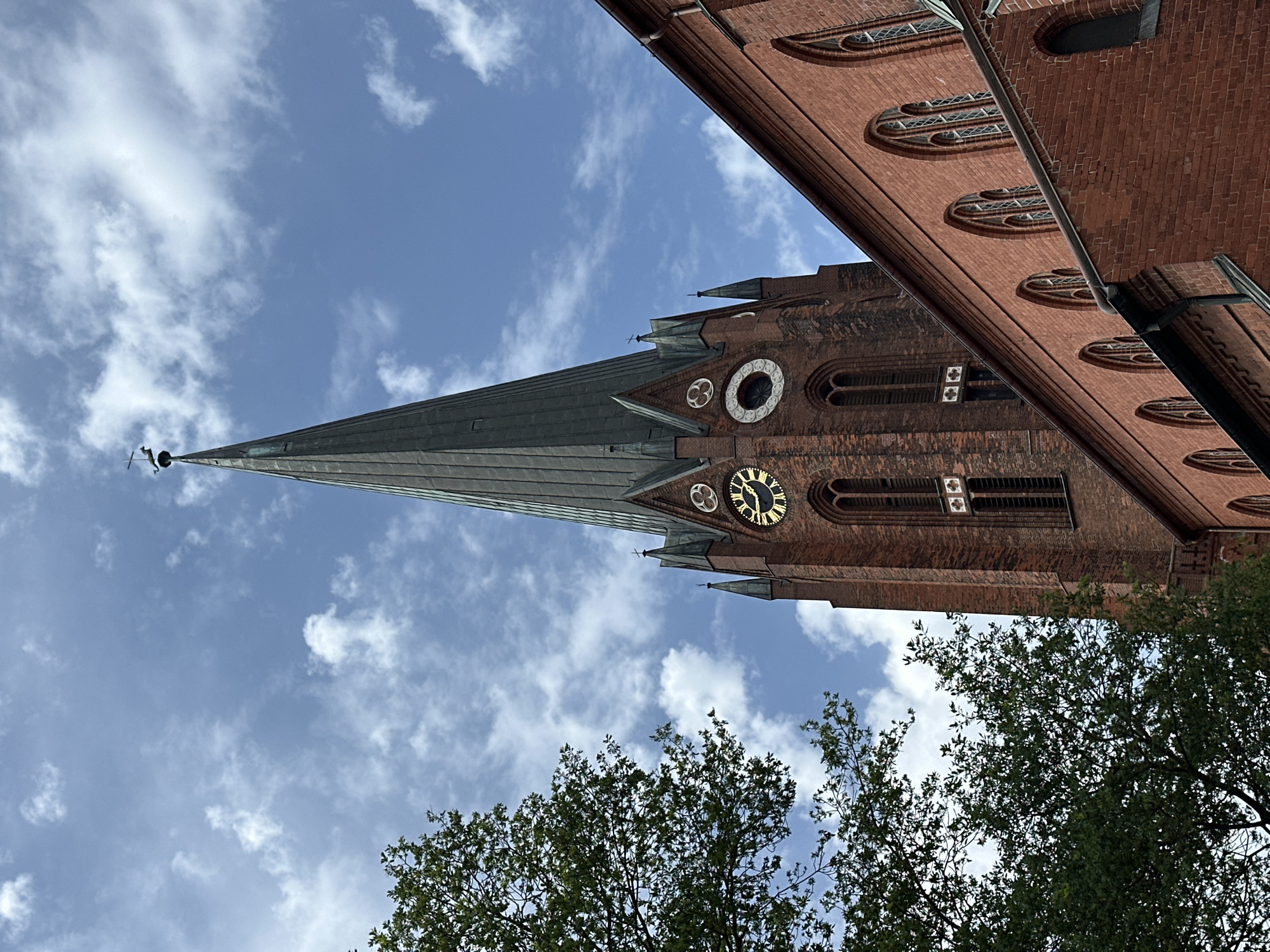 The image shows a low-angle view of the Saint Petri Church (St.-Petri-Kirche) in Buxtehude, Germany. The church's steeple rises into a partly cloudy sky. The brickwork of the church is visible, including architectural details, windows, and a clock face. Parts of the nearby trees with green leaves are visible in the foreground.