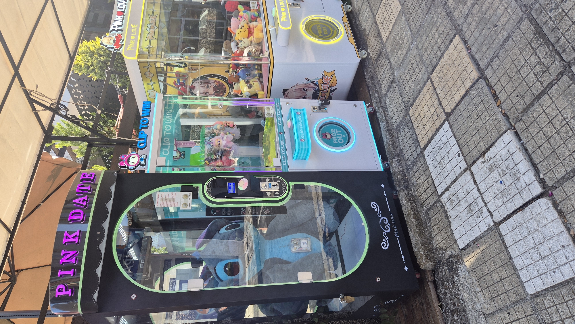 The image shows a row of three arcade claw machines parked on a tiled sidewalk in the city of Kiten, Bulgaria. The leftmost machine is black with a large glass display showing a large blue stuffed character that resembles Stitch from Lilo & Stitch. It has a sign at the top that reads "PINK DATE" in neon pink lettering. To the right of this machine is a smaller, white claw machine with a glass display filled with plush toys. This machine has a sign that says "CLIP TO WIN". The rightmost machine is also white and appears to be a similar claw machine, featuring a yellow sign with "PINK LOVE" and various plush toys inside. The machines are positioned outdoors under a light-colored awning, with trees and a building visible in the background. The ground is paved with square tiles, some of which are discolored or uneven. The scene is brightly lit by natural sunlight.