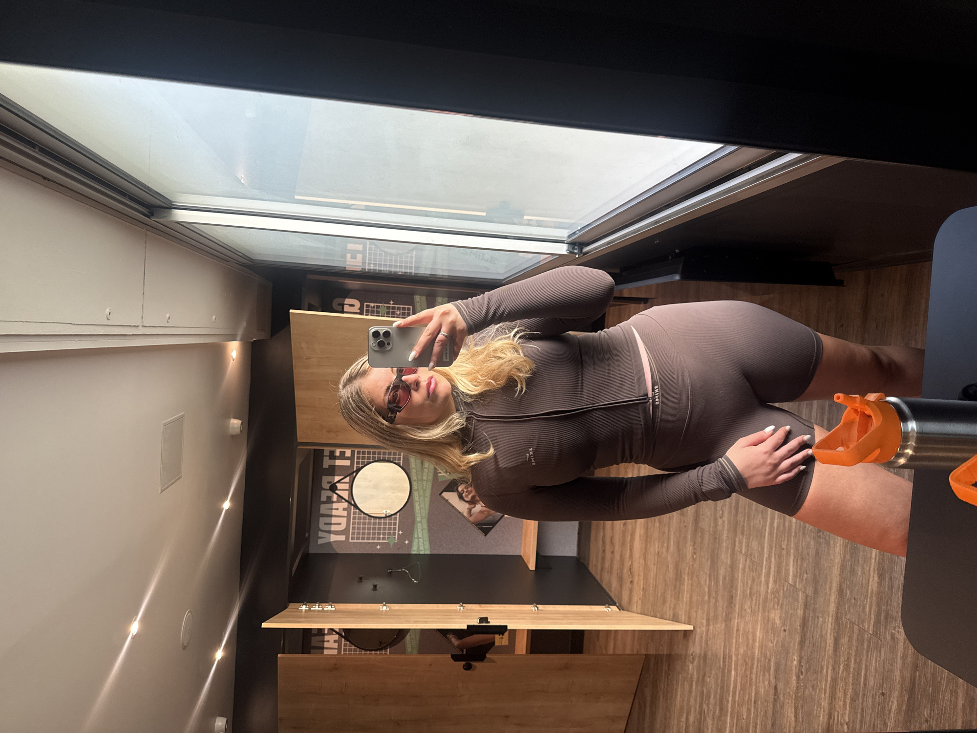 The image, rotated for proper viewing, shows a young woman taking a mirror selfie indoors. She has blonde hair, wears red-tinted sunglasses, and is dressed in a dark brown/grey ribbed athletic or leisure set, comprising a long-sleeved, high-necked top with a zipper and matching short shorts. The brand name "SKIMS" is visible on the waistband of her shorts. She holds a light-colored smartphone with a triple-lens camera array to capture her reflection.
The setting appears to be a modern indoor space, possibly a gym changing room or a retail fitting room, located in Paris, France. The floor and lower wall sections are covered in warm-toned wooden planks. To the woman's left, white cabinets or lockers with small handles are visible. To her right, part of a metal water bottle with an orange top is seen.
The background reflected in the mirror and a large overhead window or skylight reveals more details of the room. A circular mirror hangs on a dark wall behind her, next to a graphic or poster featuring reversed text that reads "EL BEADA." Further up, reflected in the window, reversed text "ICI" is faintly visible. The natural light from the window suggests it is daytime. The scene conveys a casual and contemporary interior.