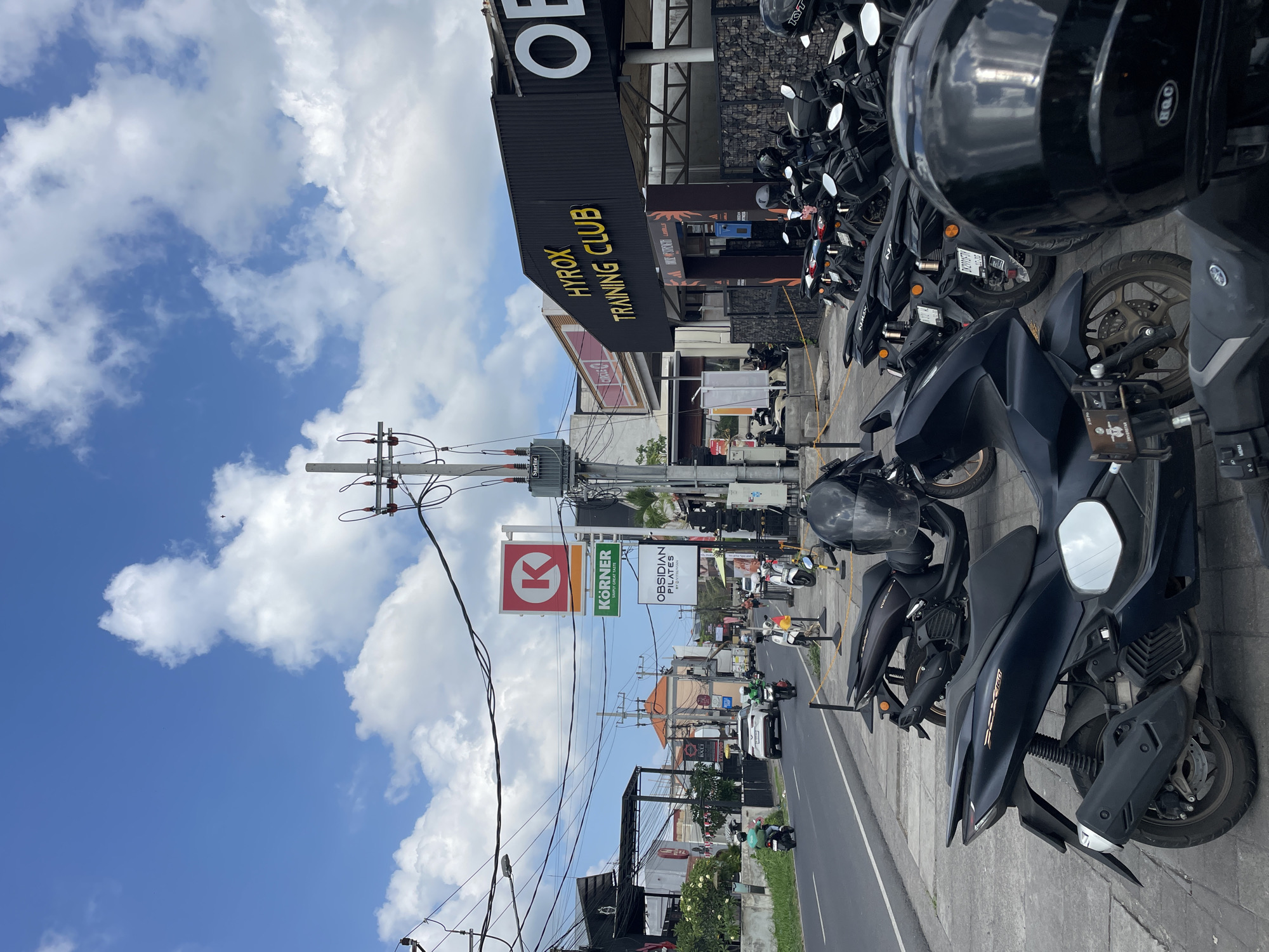 The image depicts a street scene in Mengwi, Indonesia, on a cloudy day. In the foreground, numerous parked motorcycles are lined up on a sidewalk. A black motorcycle with a silver emblem and a large black helmet is prominently featured. Further down the street, a white SUV is driving, accompanied by several other vehicles and motorcyclists.
On the right side of the street, a building with "HYROX TRAINING CLUB" written in yellow letters on a black facade is visible. Adjacent to it is another building with a red circular logo with a white "K" inside, and a green sign below it that reads "KORNER HAPPY GREAT TASTE." A sign for "OBSIDIAN PILATES" is also present.
Power lines crisscross above the street, supported by utility poles. The sky is blue with scattered white clouds. The environment appears to be an urban or commercial area, with businesses and traffic on the road.