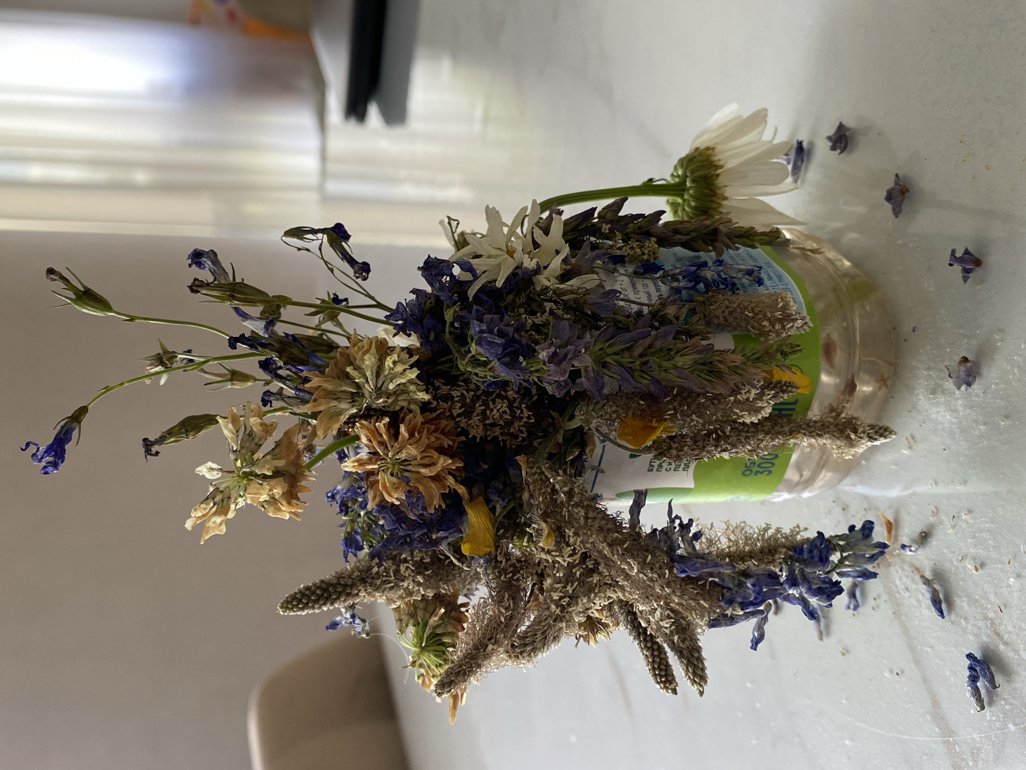 A close-up shot shows a bouquet of dried flowers in a clear plastic bottle on a white surface. The bouquet contains a variety of flowers in shades of purple, brown, and yellow, with some petals and seeds scattered on the surface. The bottle has a green label with Russian text. In the background, a blurred white curtain and part of a chair are visible, suggesting an indoor setting. The lighting is soft, casting a shadow on the left side of the image.