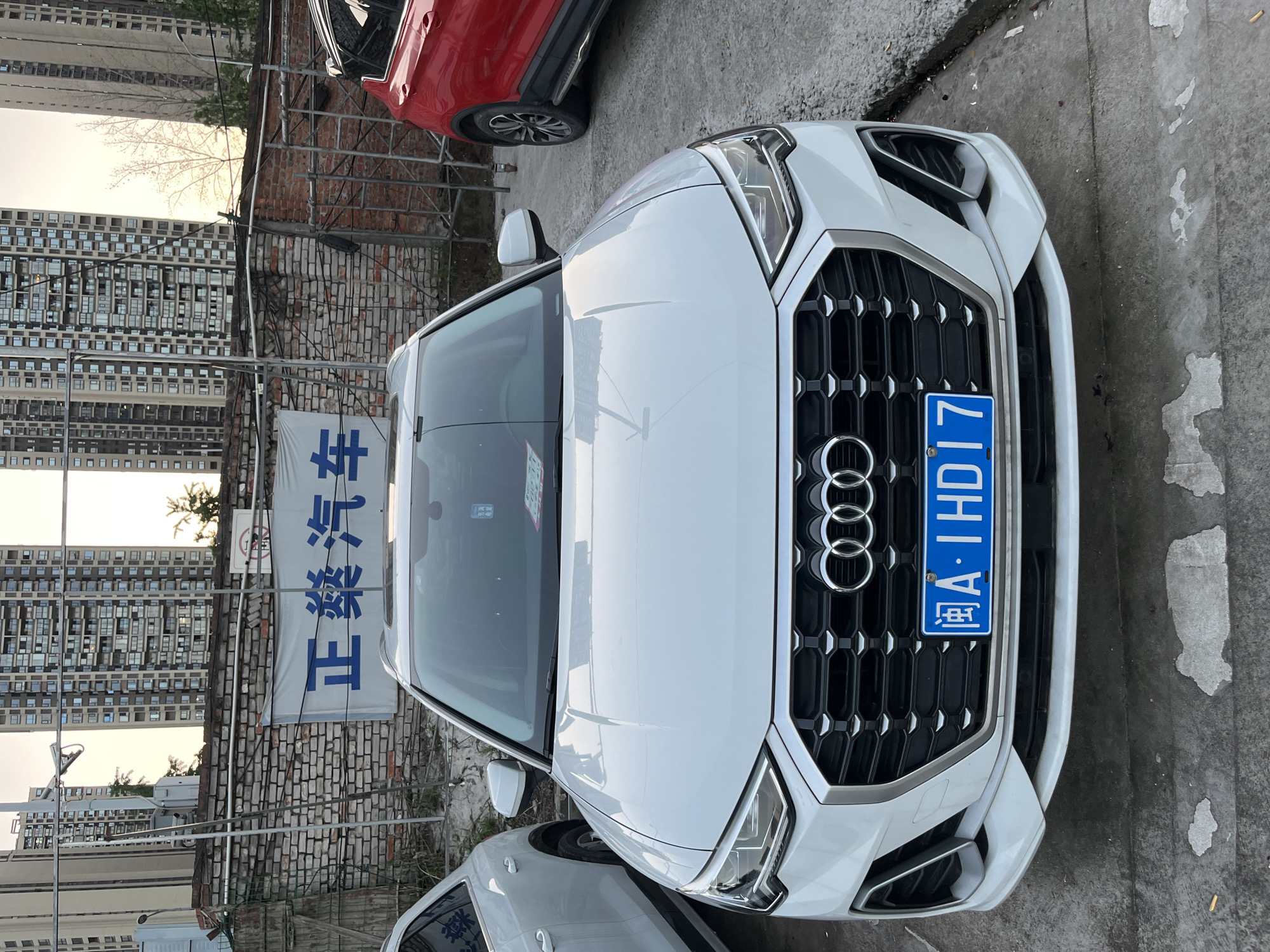 A white Audi car is parked on a paved surface. The car's license plate reads "渝A·IHD17". Behind the car, there is a brick wall, with a sign that is written in Chinese characters. In the background are tall apartment buildings. Based on the license plate, the location is likely Banan, China.