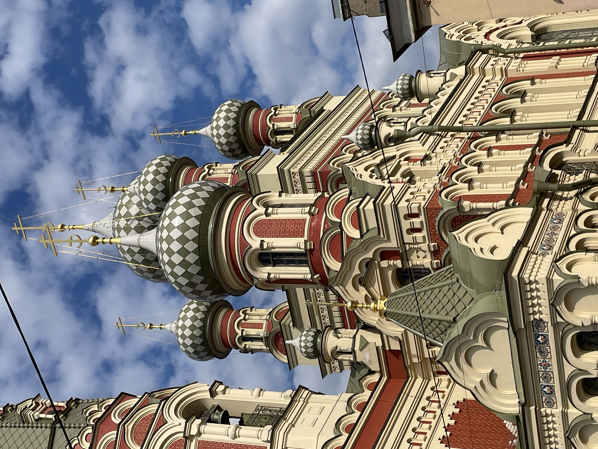 The image, correctly oriented, features a detailed, upward-angled view of an ornate Russian Orthodox church under a bright blue sky with scattered white clouds. The building is characterized by its numerous onion domes (cupolas), which are adorned with a distinctive green and white checkered pattern. Each dome is topped with a golden cross and anchor finial.
The church's facade showcases a rich array of architectural details typical of Russian Revival style. It incorporates sections of reddish-brown brickwork, contrasted with intricate cream or beige-colored elements such as pilasters, arches, and elaborate decorative moldings. Green, patterned roofs are also visible, blending with the overall design. Small, dark windows are set into the ornate walls.
The clear sky and bright lighting indicate it is daytime, likely morning or afternoon, under good weather conditions. A few black utility wires are visible against the sky and the building. Hints of other, plainer urban structures can be seen in the background on the upper right. The architectural style, particularly the patterned domes and detailed ornamentation, strongly suggests a location in Russia, consistent with Saint Petersburg. No human activity or visible text is present in the frame.