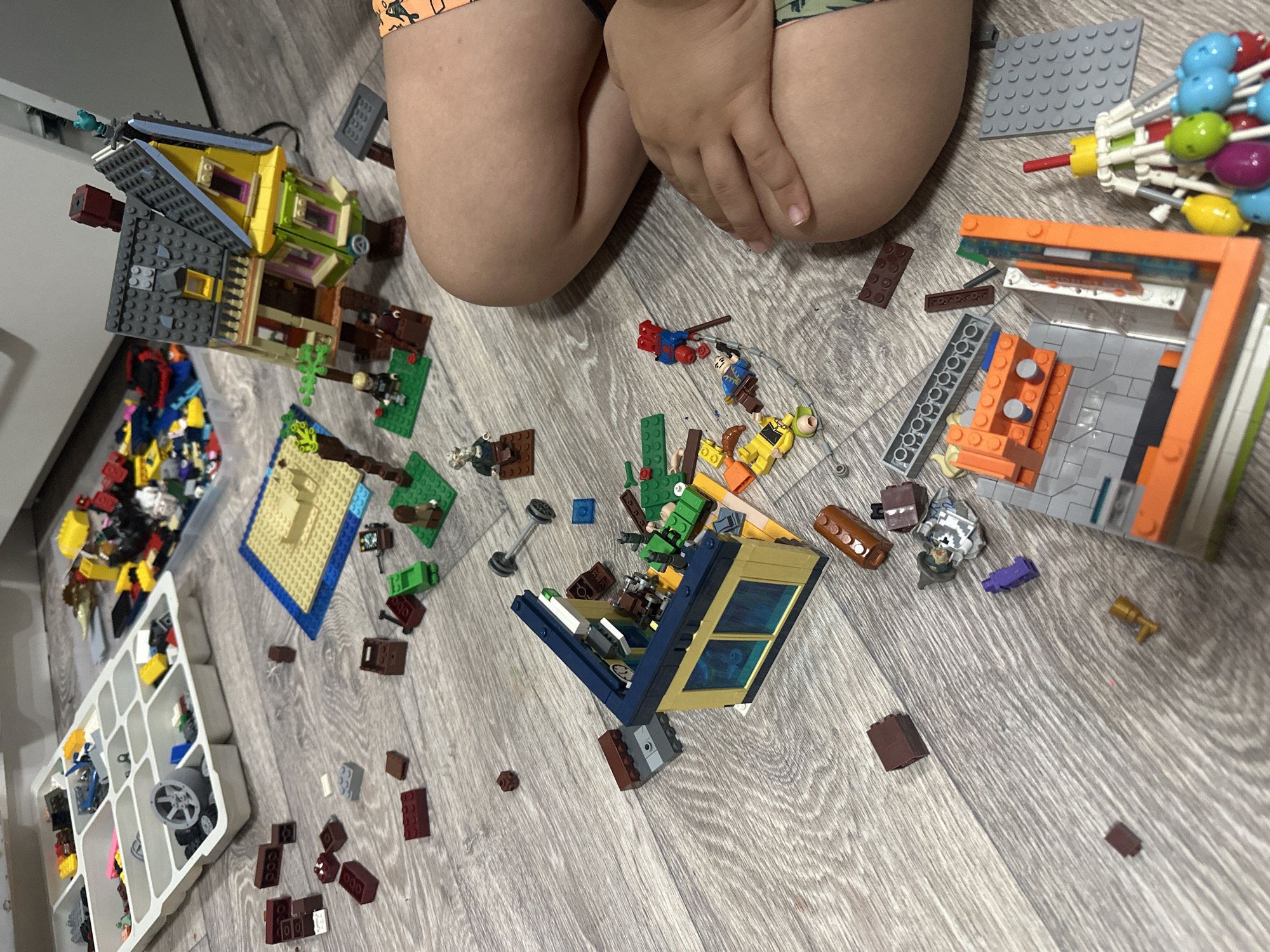 A child is kneeling on a gray wood-patterned floor, surrounded by scattered Lego bricks and partially constructed Lego sets. To the left is a plastic container holding various Lego pieces. Four distinct Lego constructions are visible: a yellow and gray house, a structure with orange and gray elements, a blue and yellow building, and a small blue and tan structure. Lego minifigures and individual Lego elements are spread across the floor.