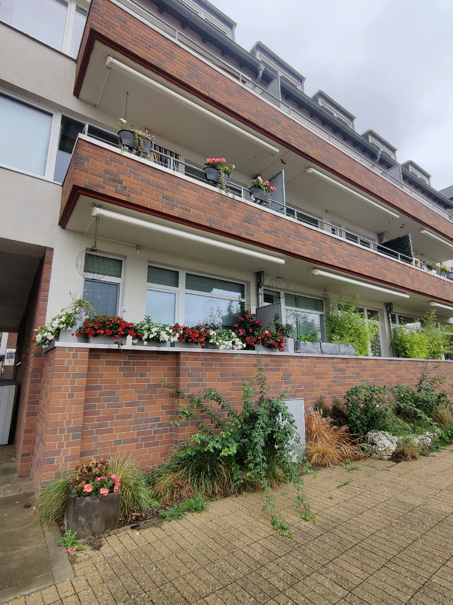 This image depicts the facade of a multi-story apartment building with brick and white siding, located in Emmerich on the Rhine, Germany. The building features multiple balconies with railings and retractable awnings. Planters filled with red and white flowers are visible on some of the balconies and along a lower brick wall. There are also several large windows and what appear to be ventilation units. The ground in front of the building is paved with tan square tiles. Greenery, including bushes and tall grasses, surrounds the base of the building and the paved area. The sky is overcast. No people are visible in the image.