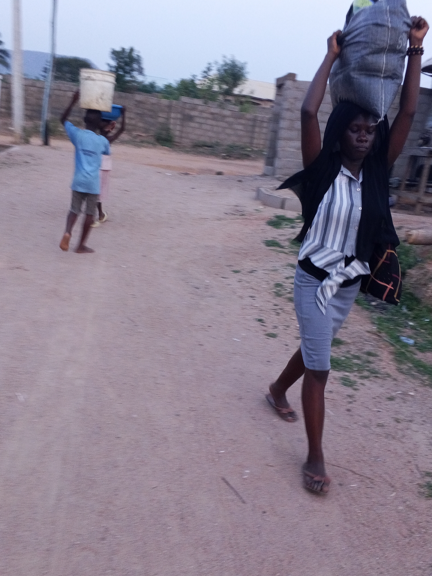 The image captures a scene in Adonkolo, Nigeria, focusing on individuals performing daily tasks. A woman is in the foreground, balancing a large sack on her head with both hands raised. She's walking towards the viewer, wearing a striped shirt, a dark-colored shawl, a knee-length skirt, and flip-flops. A patterned bag hangs from her side. In the background, two younger individuals are carrying containers on their heads as they move along the path. They are wearing light-colored shirts and shorts and are barefoot. The environment is a dirt road in what appears to be a residential area. A brick wall can be seen in the background. The sky suggests it is either early morning or late afternoon.