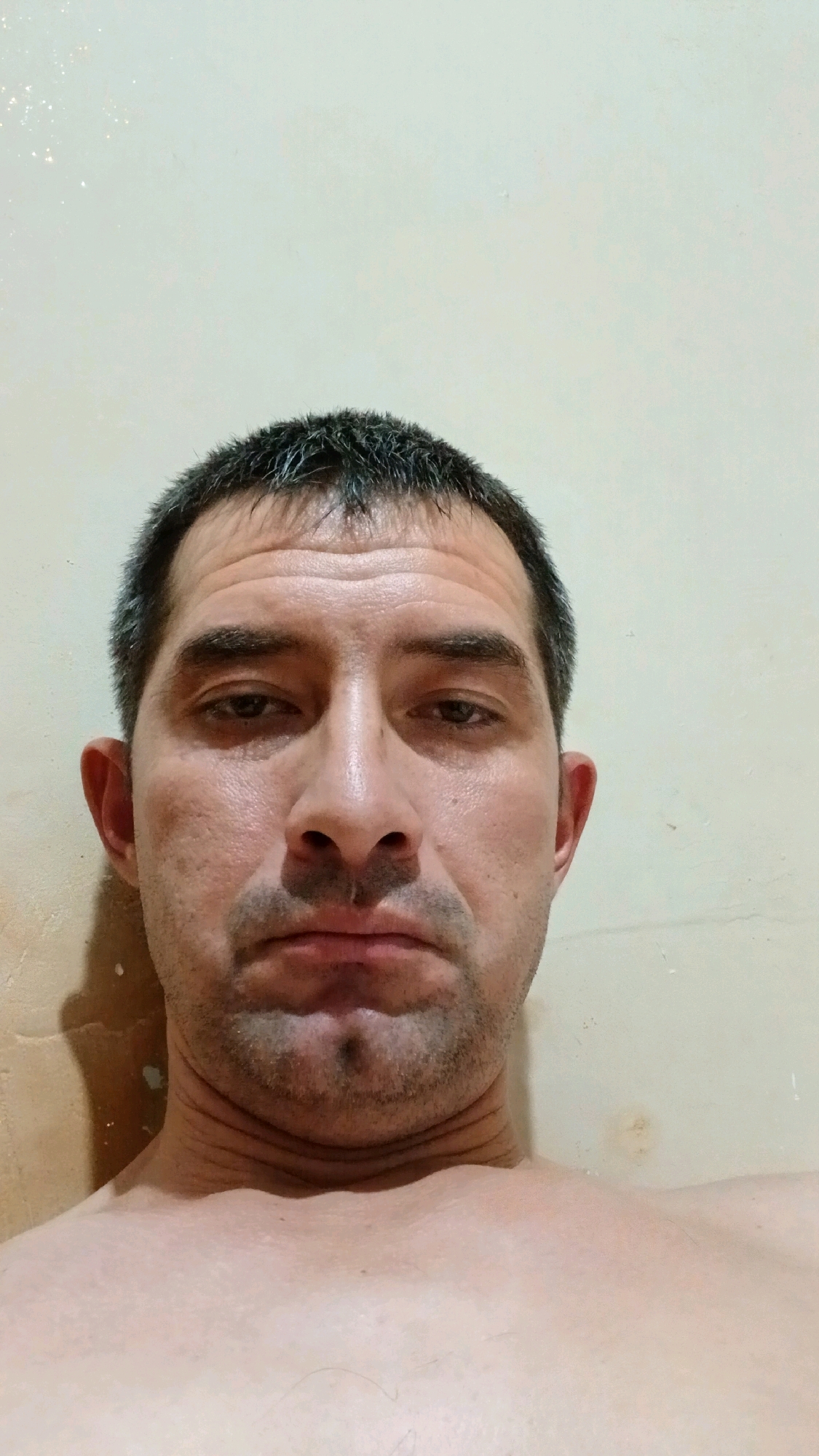 A close-up, head-and-shoulders selfie of a man is shown against a plain, light-colored wall. The man has short, dark hair with some graying. He has dark eyebrows and eyes, a prominent nose, and a mustache and goatee. His mouth is closed, and his expression appears neutral or slightly somber. His skin shows some texture and pores. He is shirtless, with his shoulders and upper chest visible. The wall behind him is textured and appears slightly dirty or stained in places, with a shadow cast on the left side of the frame. There are faint, sparkly specks on the wall, possibly from dust or reflection. The image is taken from a slightly upward angle, looking towards the man's face. The setting is indoors, with no indication of a specific location within Margilan City, Uzbekistan.