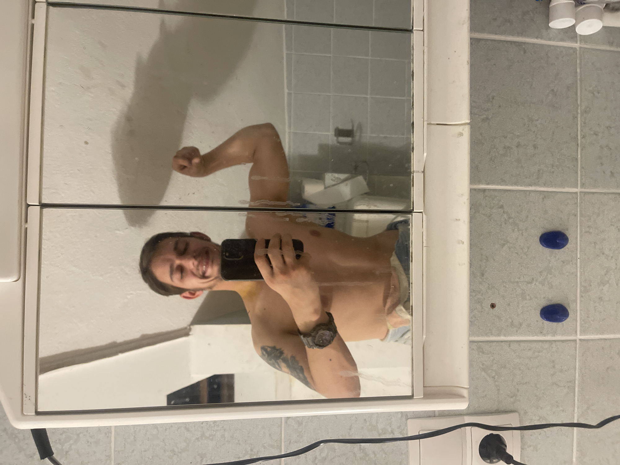 The image is a selfie taken in a bathroom mirror. A shirtless man is visible in the reflection. He is holding a black smartphone with his right hand, using it to capture the image. He is smiling. He has a tattoo on his left arm and is wearing a watch.
The bathroom features light gray tiled walls. In the background, there's a toilet and bathroom sink and accessories. A power outlet and cord are visible in the lower part of the frame. Two blue circular objects are affixed to the tiles on the right side. A small dark spot is located between them.
The location and city are not identified.