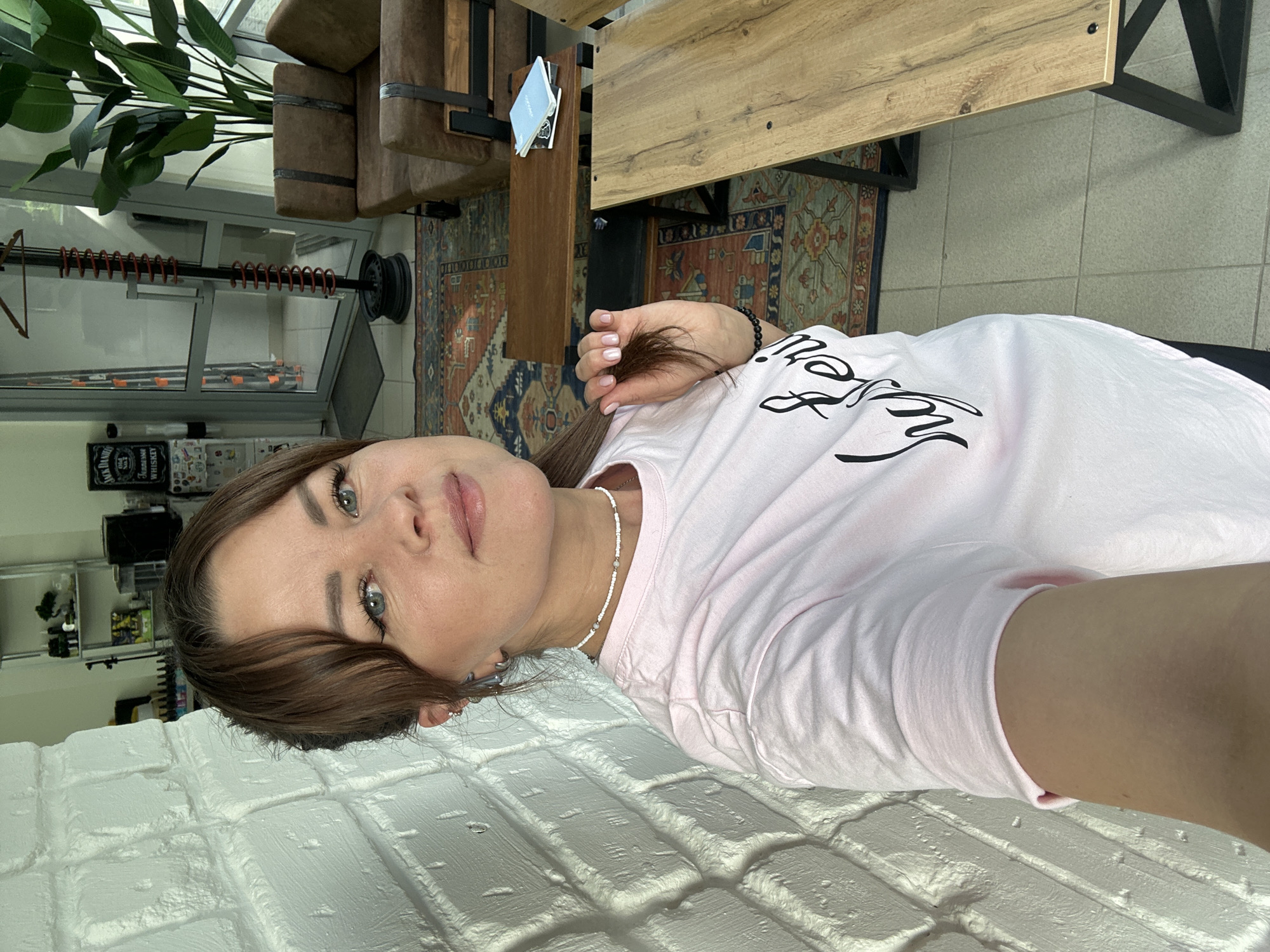 A young woman with brown hair and blue eyes is taking a selfie in what appears to be a modern, well-lit salon or waiting area. She is wearing a light pink t-shirt with black script lettering and a white beaded necklace. Her hair is pulled back, with some strands framing her face. She is leaning against a white brick wall. In the background, there are two brown leather sofas, wooden tables, a large potted plant, a glass door, and a coat rack with a spiral design. There is also a patterned rug on the floor. The overall impression is one of a clean, contemporary space. The image is not identifiable as being in Chaikovsky, Russia.