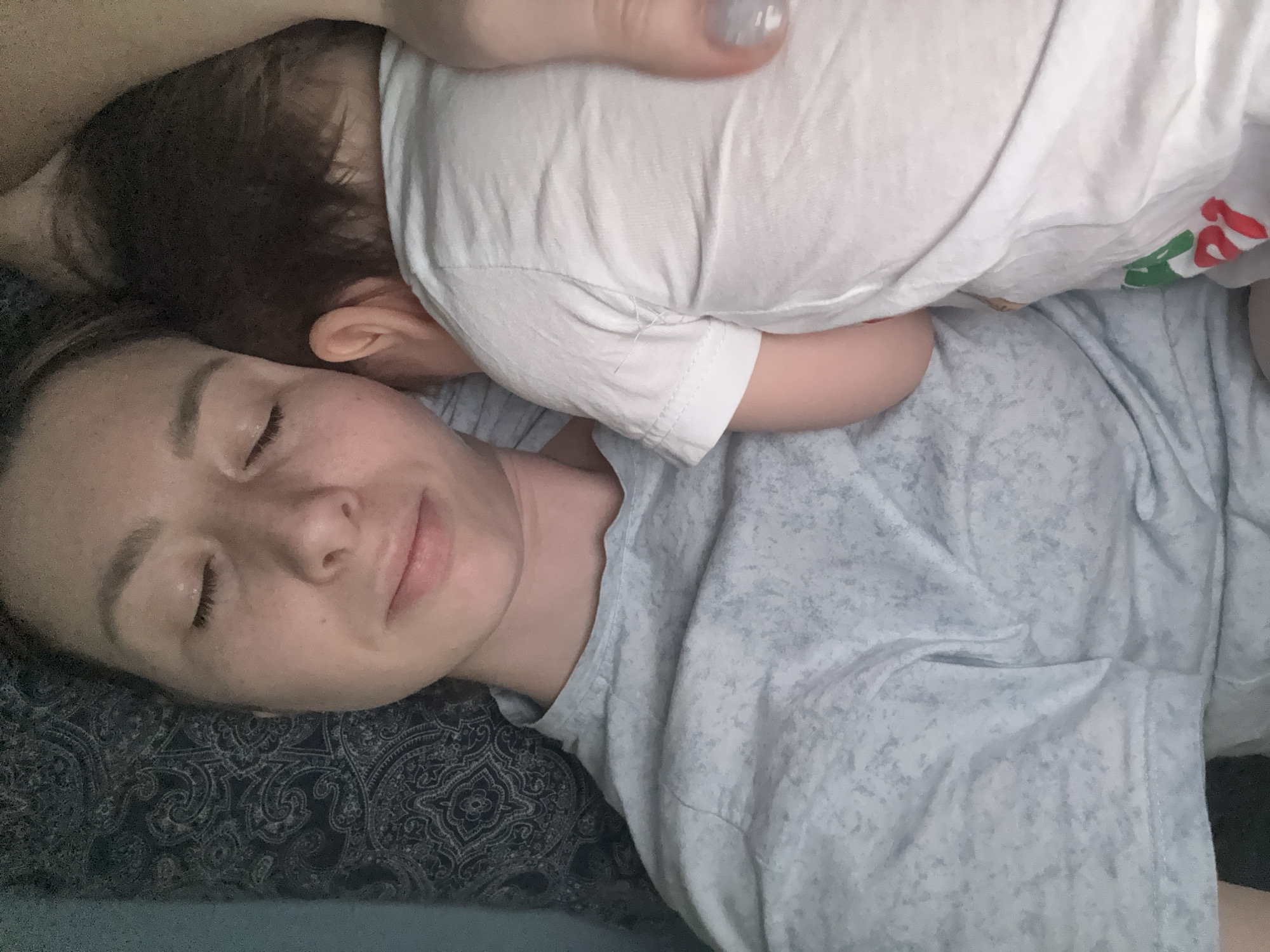 A woman is lying down with her eyes closed, and a baby is lying on top of her. The woman is wearing a light blue shirt, and the baby is wearing a white shirt. The woman is lying on a patterned pillow. The background is blurred.
The image does not contain enough information to determine the location in Moscow, Russia.