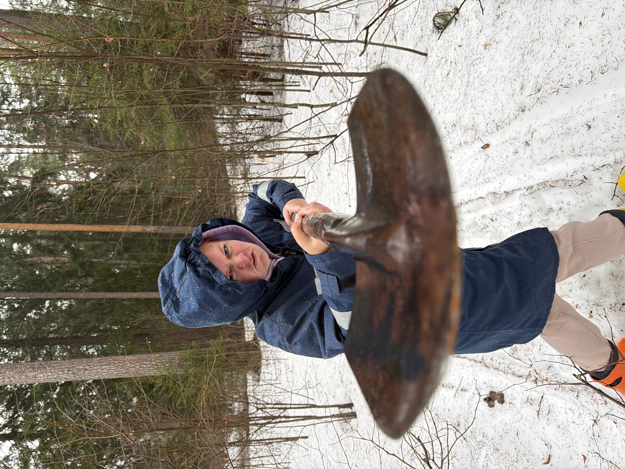 A woman wearing a blue hooded jacket, light brown pants, and orange and black boots stands in a snowy forest in Chaikovsky, Russia. She is holding a rusty shovel in front of her, obscuring part of the view. The ground is covered in snow and scattered with small twigs and leaves. Bare trees are visible in the background.