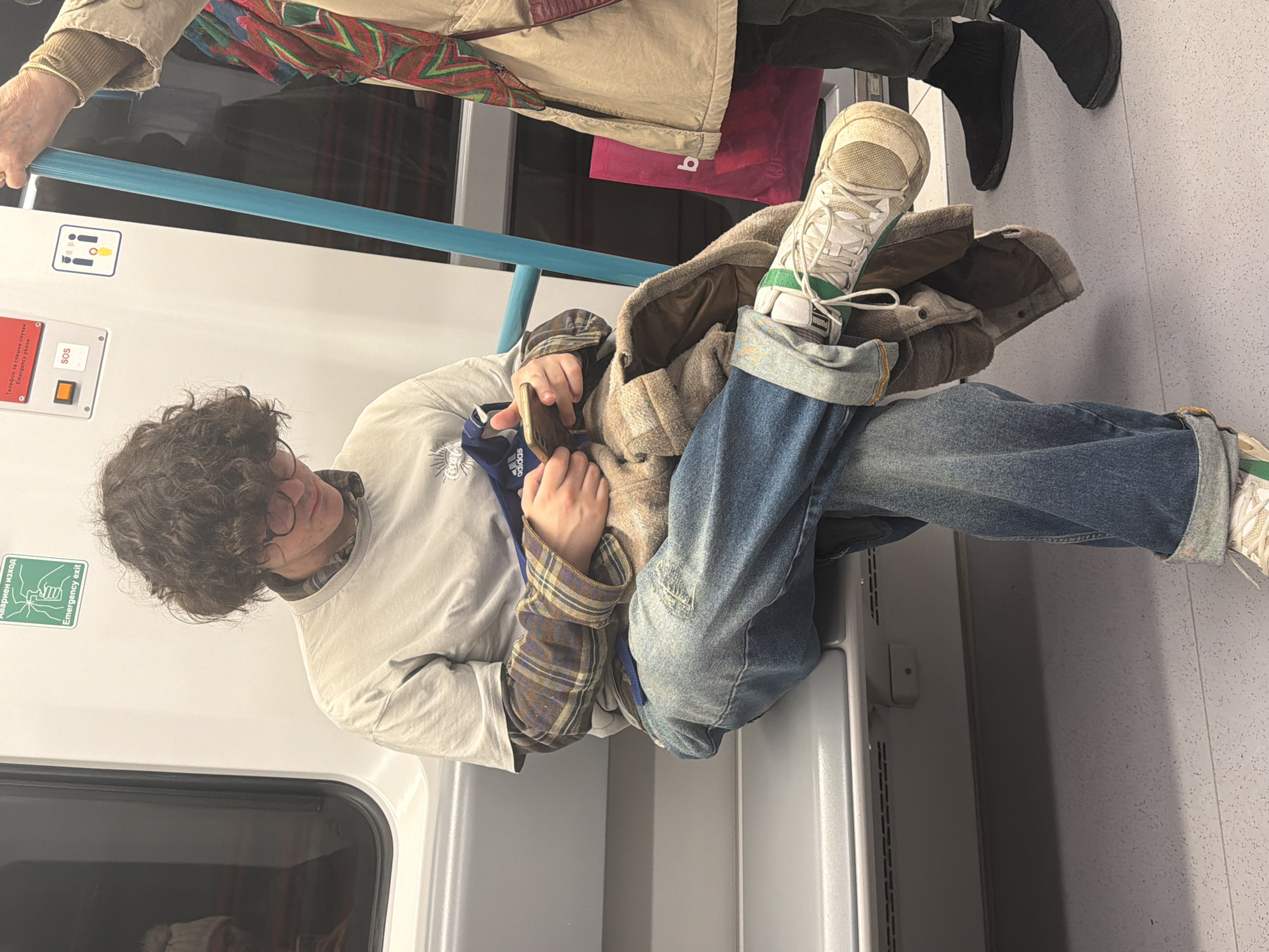 The image shows a young person sitting on a seat in what appears to be a subway car. The person has curly hair, is wearing glasses, a white t-shirt, a plaid shirt and blue jeans. They are holding a phone. Another passenger is visible in the background. The environment is a subway train car with white walls, teal handrails, and informational signage. Based on the signage which is written in Cyrillic and the emergency exit green box writing on the wall, the location is likely within the city of Sofia, Bulgaria.