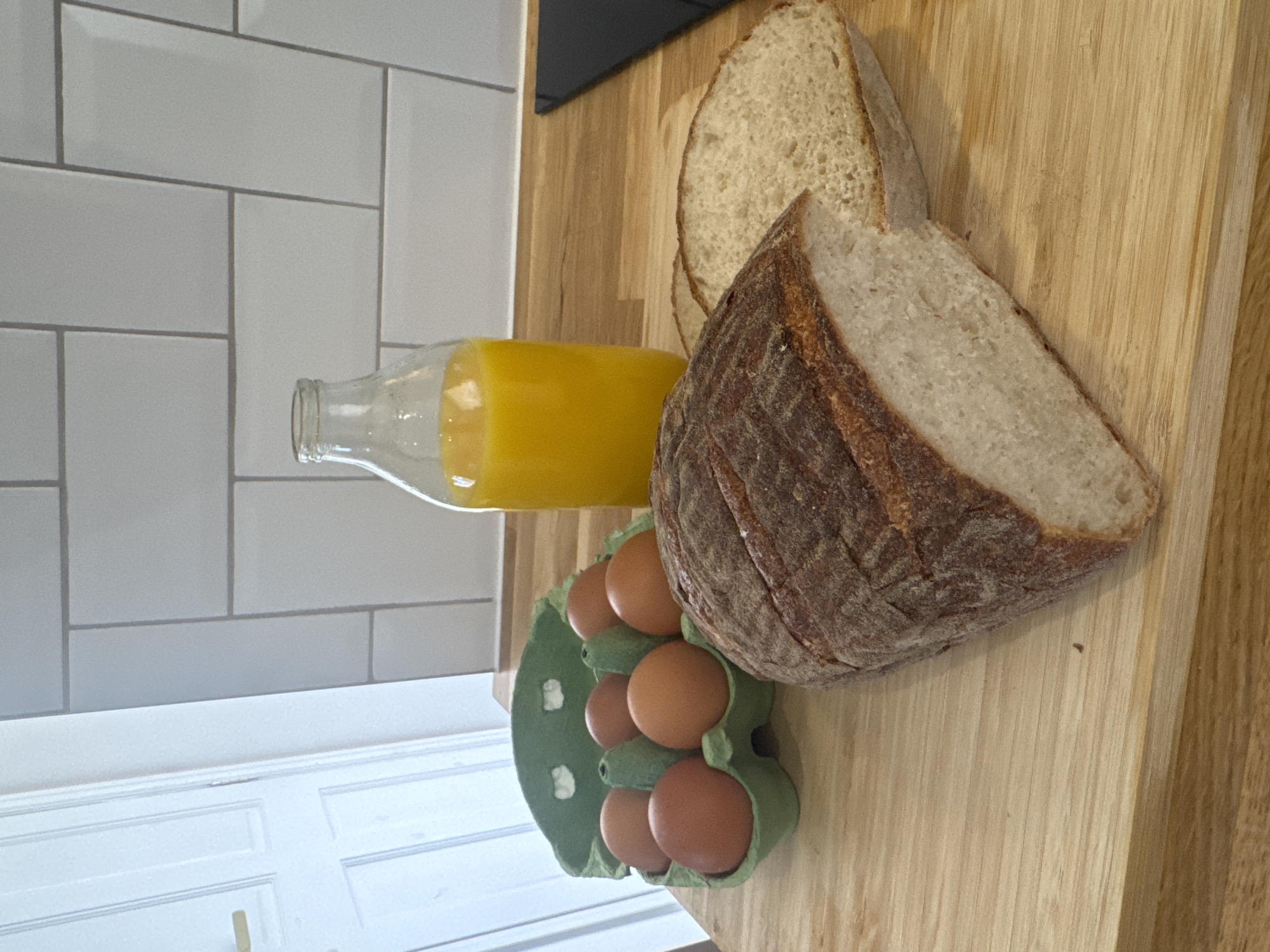 A still life image shows a breakfast spread on a light-colored wooden countertop. In the foreground, a rustic loaf of bread is partially sliced, revealing its white, airy interior and dark crust. To the left of the bread, a green egg carton holds six brown eggs. Behind the eggs, a clear glass bottle is filled with bright orange juice. The background features a tiled wall with white rectangular tiles laid in a brick pattern. To the far left, a portion of a white door with a gold handle is visible. The scene is lit from above, casting soft shadows. The overall impression is one of a simple, wholesome breakfast preparation.