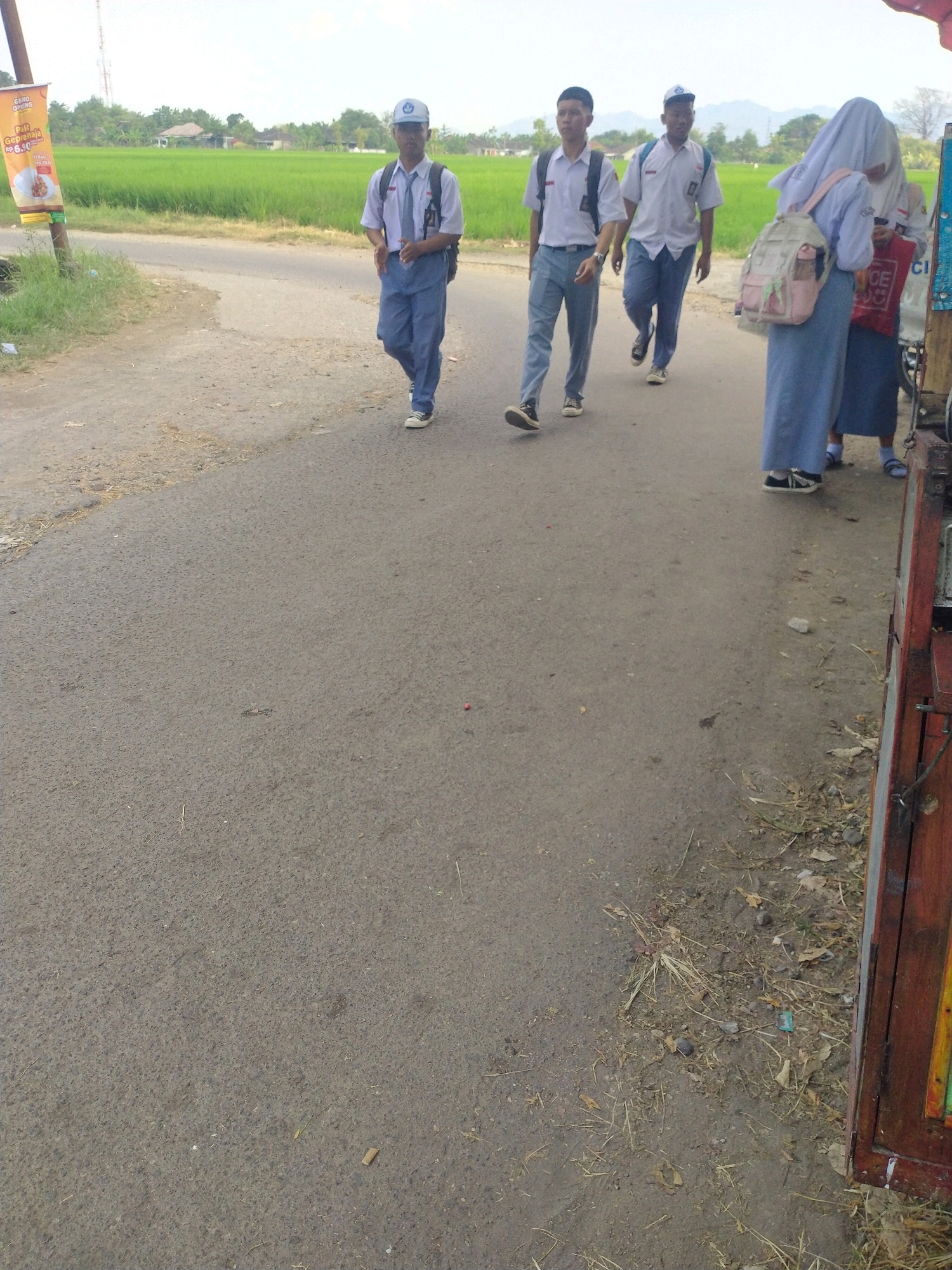 In a rural setting near the city of Begajah, Indonesia, four individuals, presumed to be students, are captured walking on a paved road. The image is dominated by the road surface, which shows signs of wear and tear, with scattered debris including dry grass and small stones. To the left of the road, a vibrant green rice field stretches towards the horizon, bordered by scattered trees and low-lying buildings under a bright, overcast sky. A prominent advertising pole stands near the road, featuring an orange banner with Indonesian text and images of food.
The four individuals are walking in the same direction, away from the viewer. The foremost person on the left is a male wearing a school uniform consisting of a white short-sleeved shirt, blue pants, a blue tie, and a white cap with a blue emblem. He has a backpack on. To his right, a second male student is also in uniform: a white short-sleeved shirt with a blue tie and a blue baseball cap. He is wearing dark pants and carrying a backpack. Further to the right, a third student, also male, is dressed in a similar school uniform, including a white short-sleeved shirt and dark pants. He is wearing a light blue backpack. The last individual on the far right appears to be a female student, her face obscured by a white veil that also covers her head. She is wearing a blue skirt and a light-colored backpack with pink accents. Her attire suggests she is also in school uniform.
The right edge of the image is occupied by the side of a wooden structure, possibly a stall or cart, painted with colorful panels, including yellow, green, and red. The overall atmosphere is one of everyday life in a semi-urban or rural Indonesian community, likely during the daytime given the lighting.