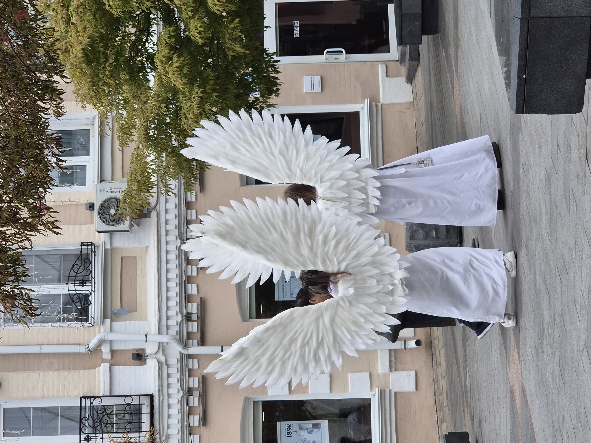 Two individuals wearing large white angel wings and white robes stand facing away from the camera on a paved walkway in front of a building. The building has a pale peach facade with white decorative trim and windows with black wrought iron balconies. Trees with green and brown leaves are visible behind the individuals and the building. One individual on the left has dark hair tied in a ponytail. The individual on the right's face is obscured. The ground is paved with grey tiles with a patterned inlay. The overall scene suggests an outdoor event or performance in a city environment. The provided hints indicate the location as Vladivostok, Russia, based on Cyrillic text on signage in the building's windows, specifically "АМЕРИКАНО" and "РЕЖИМ РАБОТЫ".