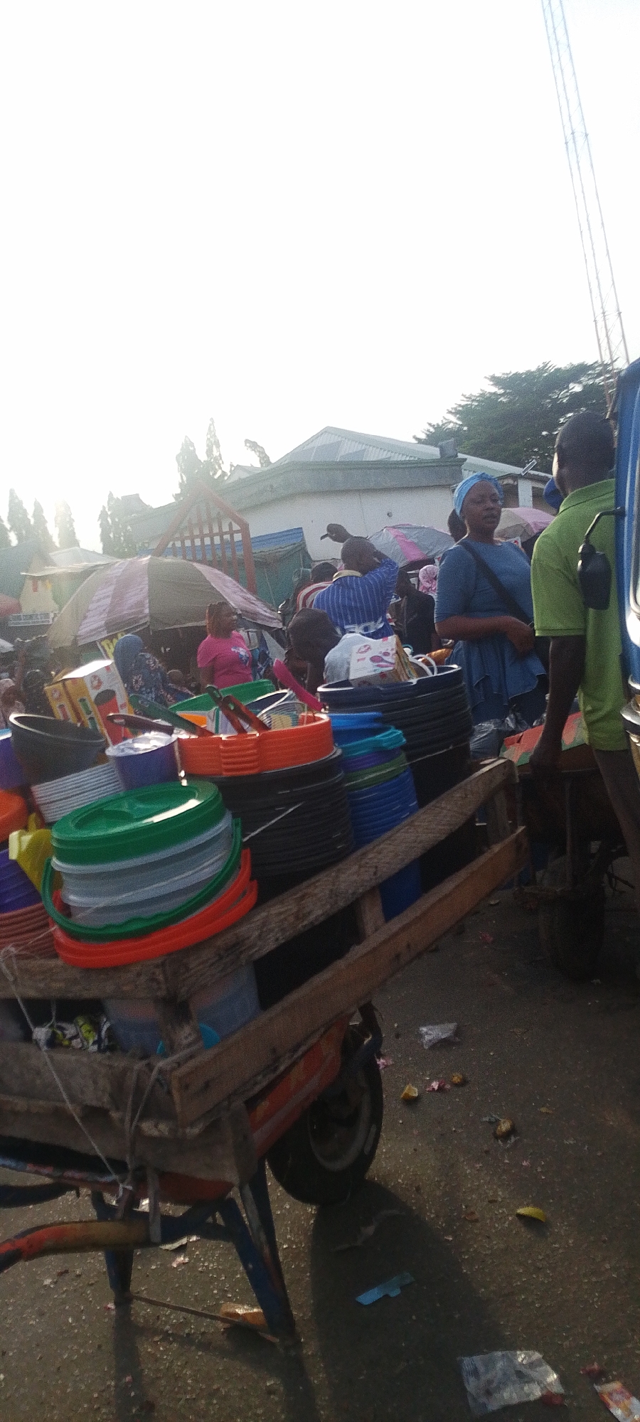 The image captures a vibrant, sunlit outdoor market scene, likely in Lokoja, Nigeria, based on the provided context. The setting appears busy, with numerous people and market stalls.
In the foreground, a sturdy wooden cart, possibly a tricycle converted for cargo, is laden with an abundance of colorful plastic household items. Stacks of buckets and basins in shades of green, orange, red, black, blue, and white are prominently displayed. Other items like strainers and storage containers are also visible among the merchandise, suggesting a vendor specializing in kitchenware or general household plastics. The cart's wheels and structure show signs of use, with painted blue and red accents.
In the mid-ground, several individuals are engaged in market activities. On the right, a man wearing a lime green polo shirt is seen from behind, standing next to the cart, possibly a vendor or helper. He has a black object clipped to his side. To his left, a woman in a blue dress and a light blue headscarf stands, looking towards the left, appearing to interact with someone or observe the surroundings. Further into the market, under various colorful umbrellas providing shade, other people are visible, including a woman in a pink shirt.
The background shows more market activity with various stalls and structures. Trees and buildings are discernible, some with sloped roofs, under a bright, hazy sky. The sun, likely either morning or late afternoon, casts soft, elongated shadows on the ground from the cart and people, indicating its low angle. The ground is an unpaved surface, with some scattered debris and litter, typical of a bustling market environment. One of the cropped images reveals a tall communication tower in the distant background, though it's largely obscured in the main image. No discernible text is clearly visible on any signs or objects.