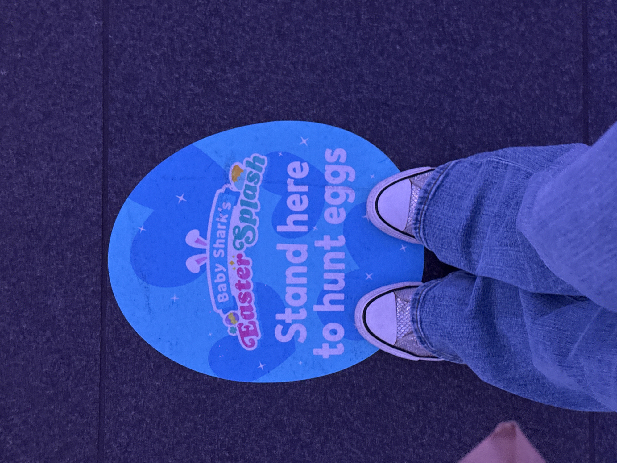 A blue oval sign on a dark grey floor reads "Baby Shark's Easter Splash. Stand here to hunt eggs." A person wearing blue jeans and white and silver sneakers is standing on the sign. The location is likely the London Aquarium, United Kingdom.