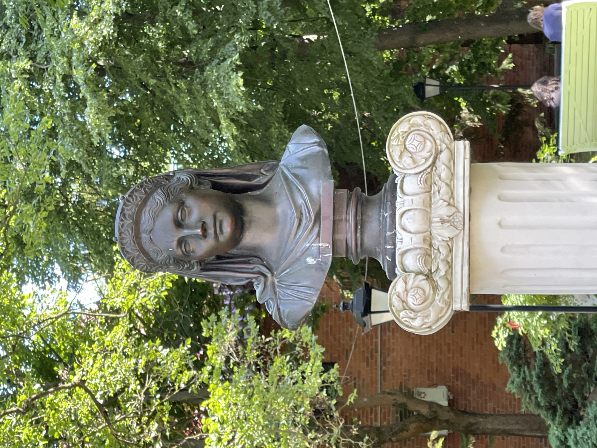 The media file shows a close-up, vertical shot of a bronze bust of a woman, displayed outdoors. The bust is positioned in front of a white fluted column with ornate carvings, featuring floral motifs. Behind the bust and column, a dense canopy of green trees with dappled sunlight filtering through suggests a daytime setting. In the background, a brick wall is visible, along with a black lamppost. To the far right, a portion of a green bench and some people are visible, one person wearing a blue top. The overall impression is one of a peaceful, possibly park-like, outdoor environment. The bust itself has a serene expression.