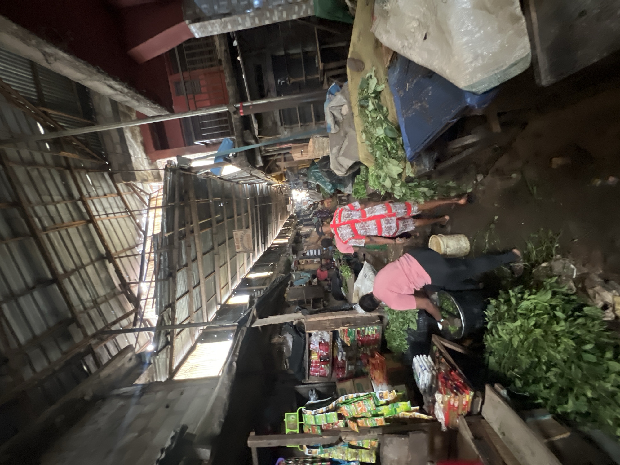 The image depicts a long, narrow market scene in Nkpor, Nigeria. The market is covered by a patchwork of corrugated metal and wooden structures forming a dimly lit corridor. The ground appears muddy or wet in places. People are visible throughout the market, some bending over goods or walking along the aisle. Stalls line both sides of the market corridor, displaying a variety of items including packaged goods, fresh produce (specifically leafy greens), and other merchandise. The stalls are constructed from rough wooden planks and metal supports. The overall atmosphere is one of bustling commerce in a rudimentary infrastructure setting.