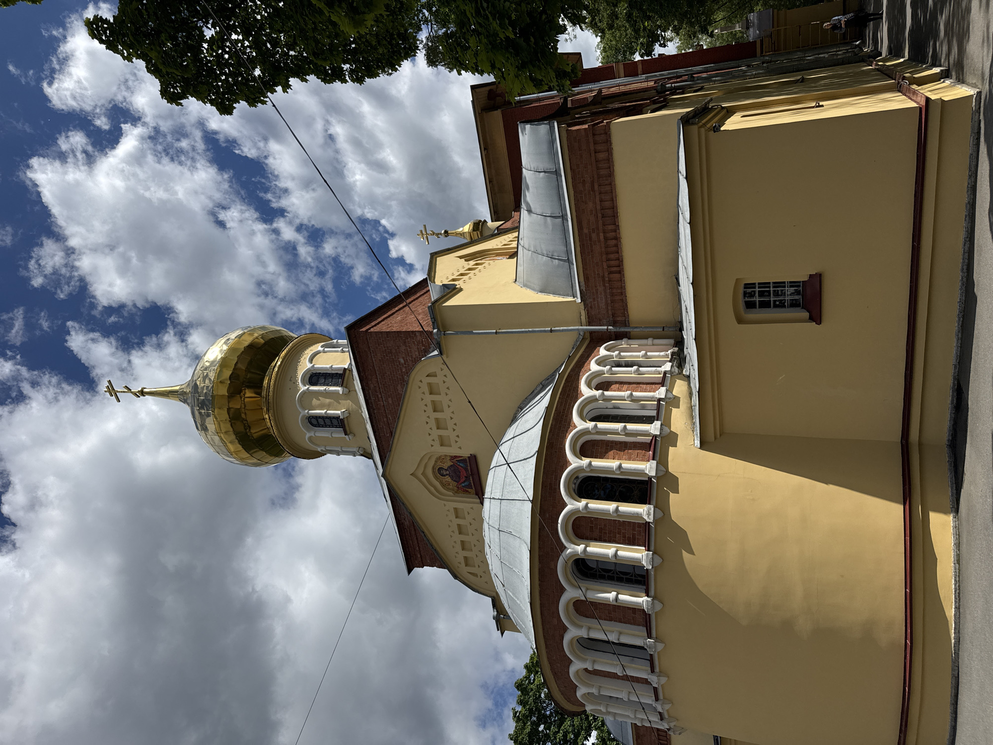 The image displays the exterior of a yellow church with a gold dome and a cross on top. The church has a red brick facade around the windows and along the roofline. The windows are arched and have a white decorative trim. There is a mosaic of a religious figure above one of the arched windows. The sky is blue with white, fluffy clouds. Trees with green leaves are visible at the top of the image. The sunlight casts shadows on the side of the building, suggesting it is daytime. A thin black wire runs diagonally across the sky. The scene is a view of a church in what appears to be a temperate climate. There is no visible text in the image.